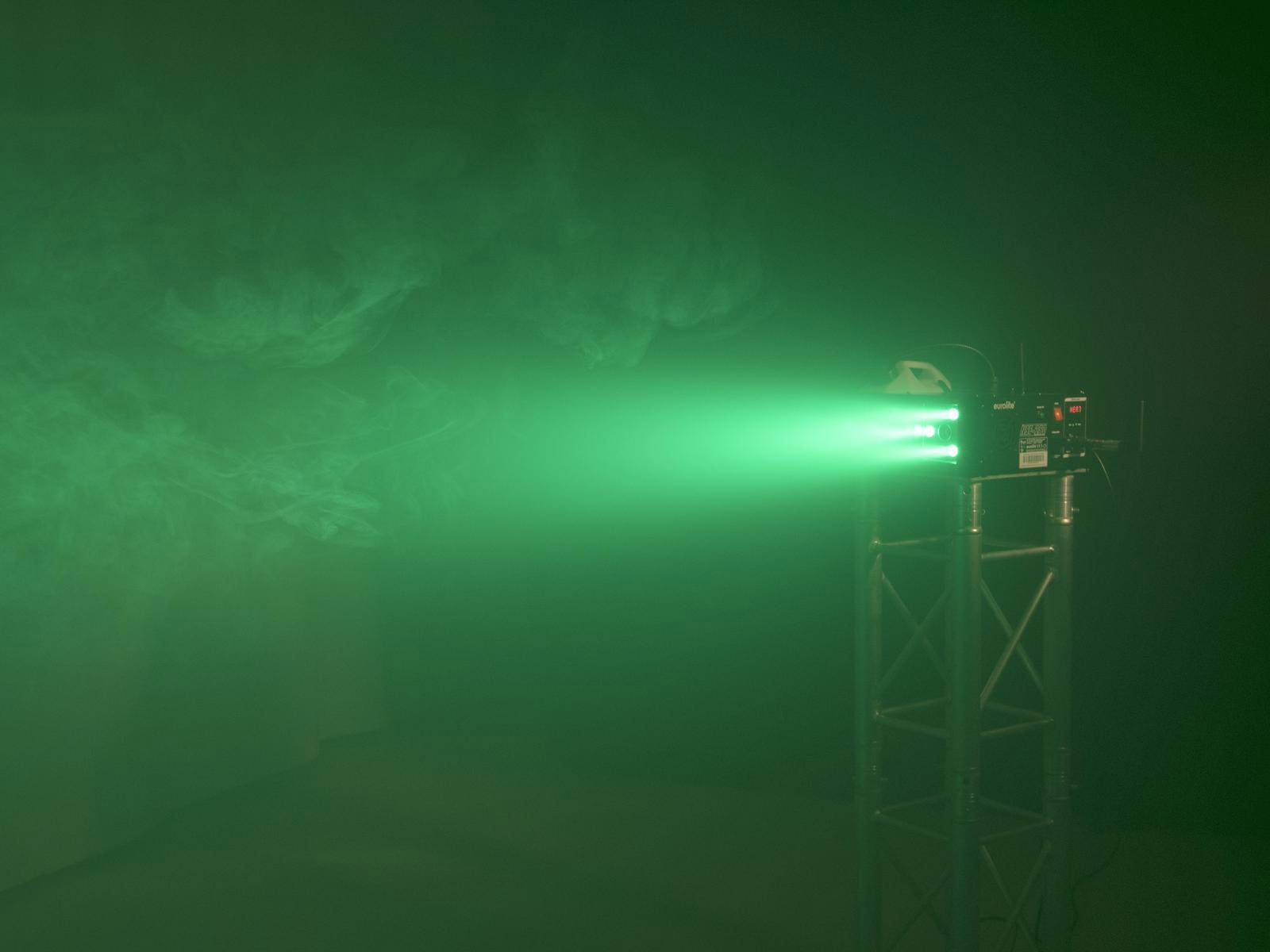Ein grüner Laserstrahl wird in einem dunklen Raum projiziert, der von Nebel durchzogen ist. Die Lichtquelle steht auf einem Metallgestell.