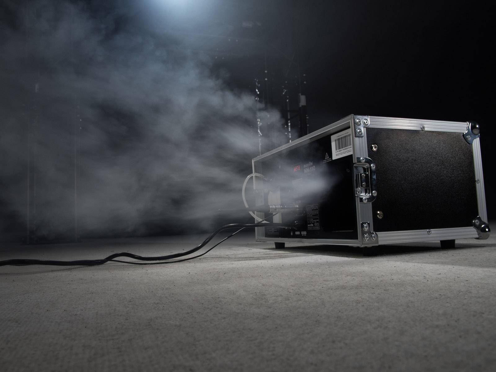 Ein Nebelmaschine auf einem Boden erzeugt dichten weißen Rauch. Kabel sind angeschlossen. Die Szene ist schwach beleuchtet.