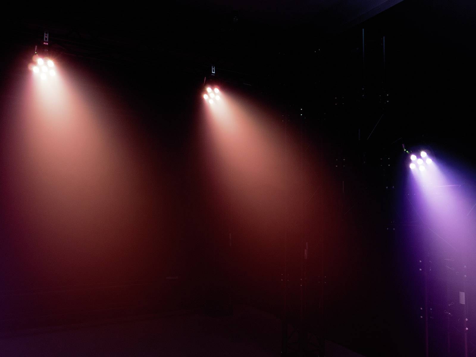 Drei bunte Scheinwerfer leuchten auf eine dunkle Bühne. Links sind sie rot und lila, rechts violett. Keine Personen sichtbar.