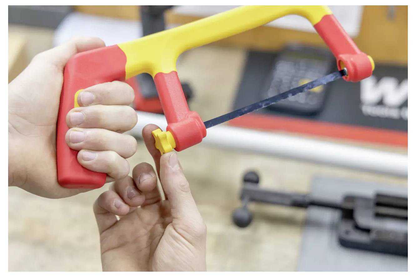 Eine Person stellt die Spannung einer kleinen tragbaren Handsäge mit roten und gelben Griffen ein, die auf einer Werkbank mit unscharfen Werkzeugen im Hintergrund liegt.