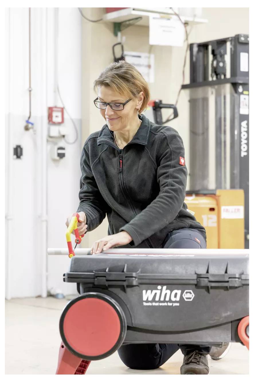 Eine Person, die eine Jacke trägt, benutzt ein Werkzeug auf einer Werkbank mit Rädern. Industrielle Ausrüstung und ein Feueralarm sind im Hintergrund sichtbar.