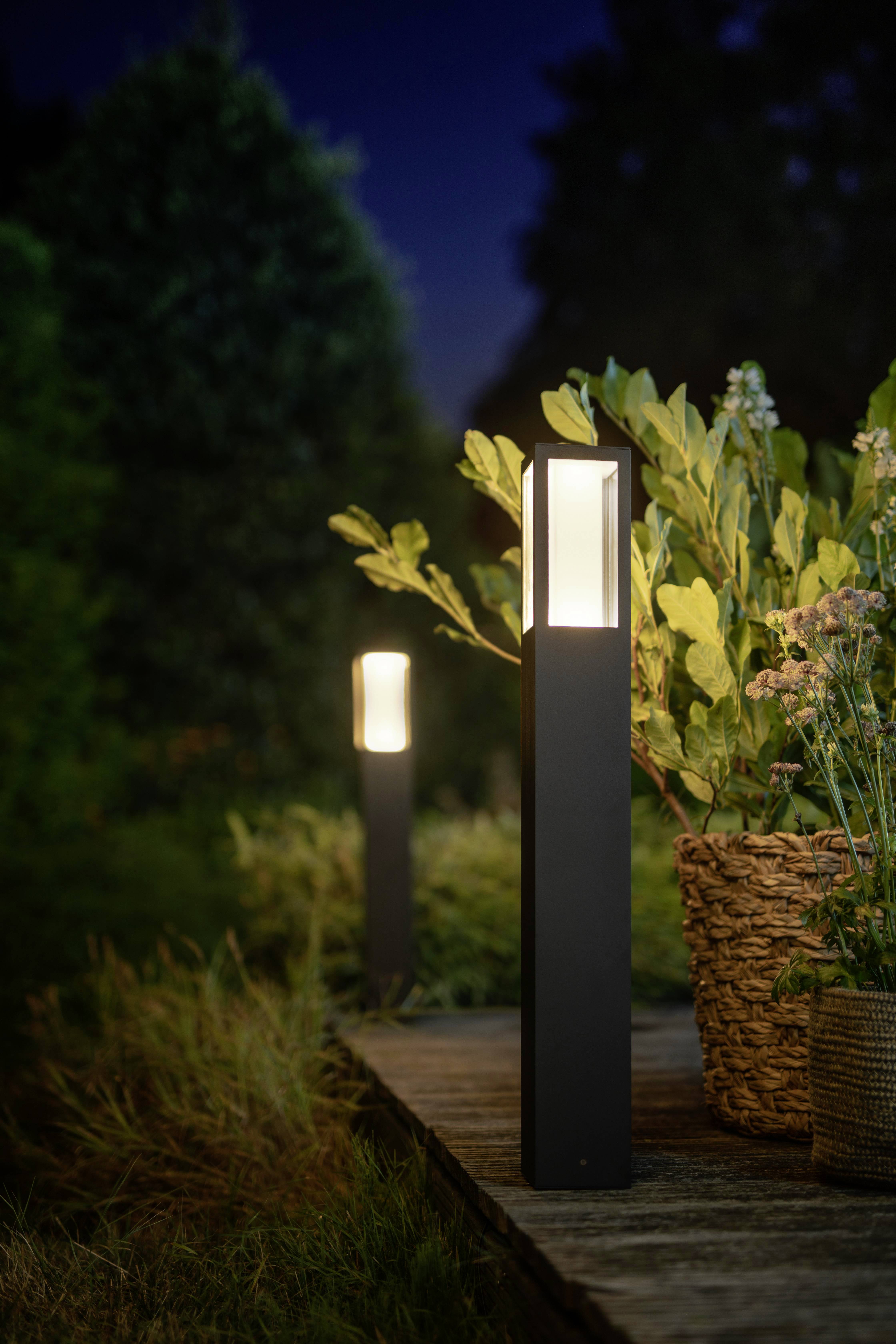 Zwei moderne, leuchtende Gartenlampen beleuchten einen Gehweg bei Nacht. Pflanzen und Blumen stehen neben den Lampen.