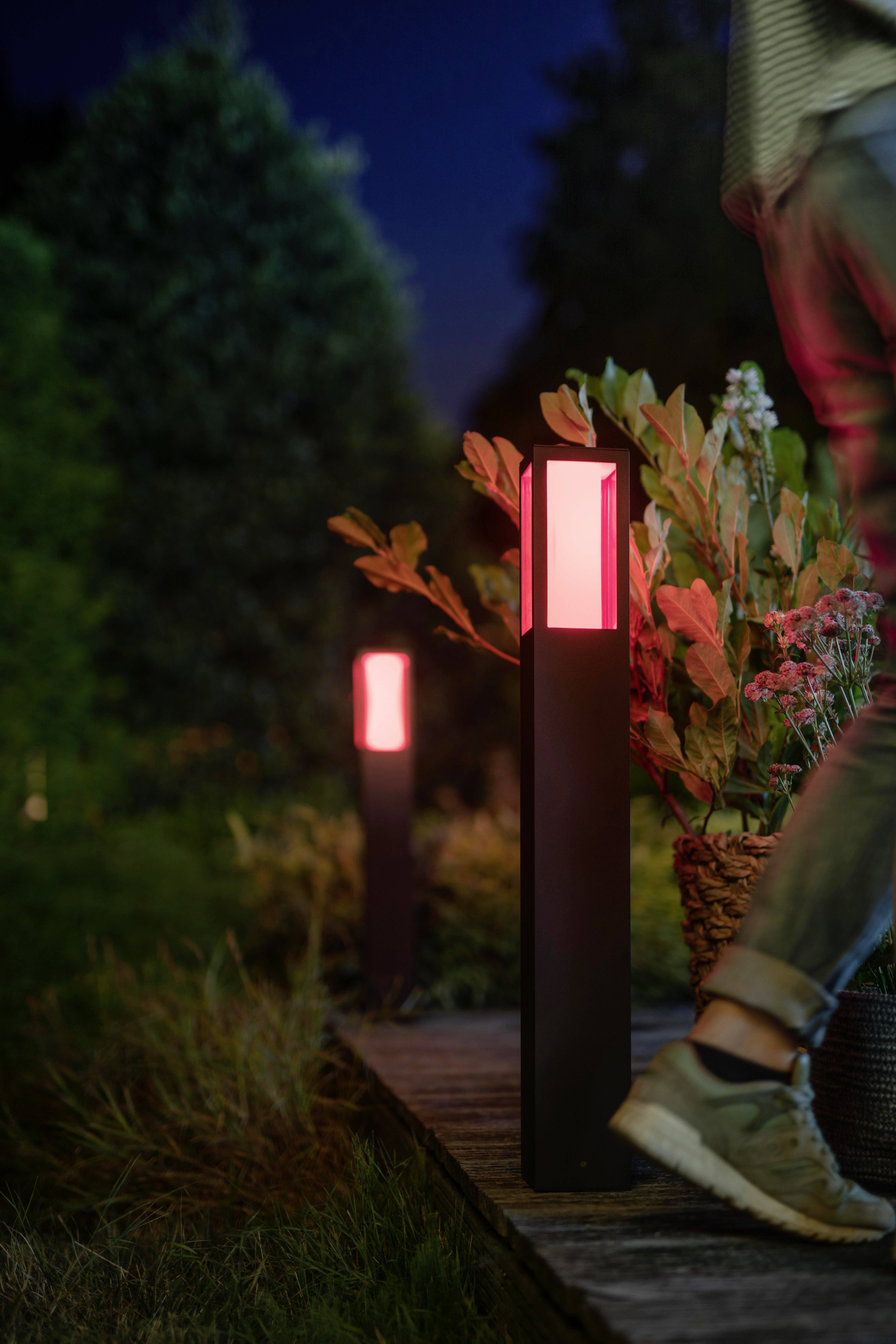 Eine Person geht nachts auf einem Holzweg entlang, flankiert von modernen Gartenlampen, die ein rotes Licht ausstrahlen, umgeben von Pflanzen.