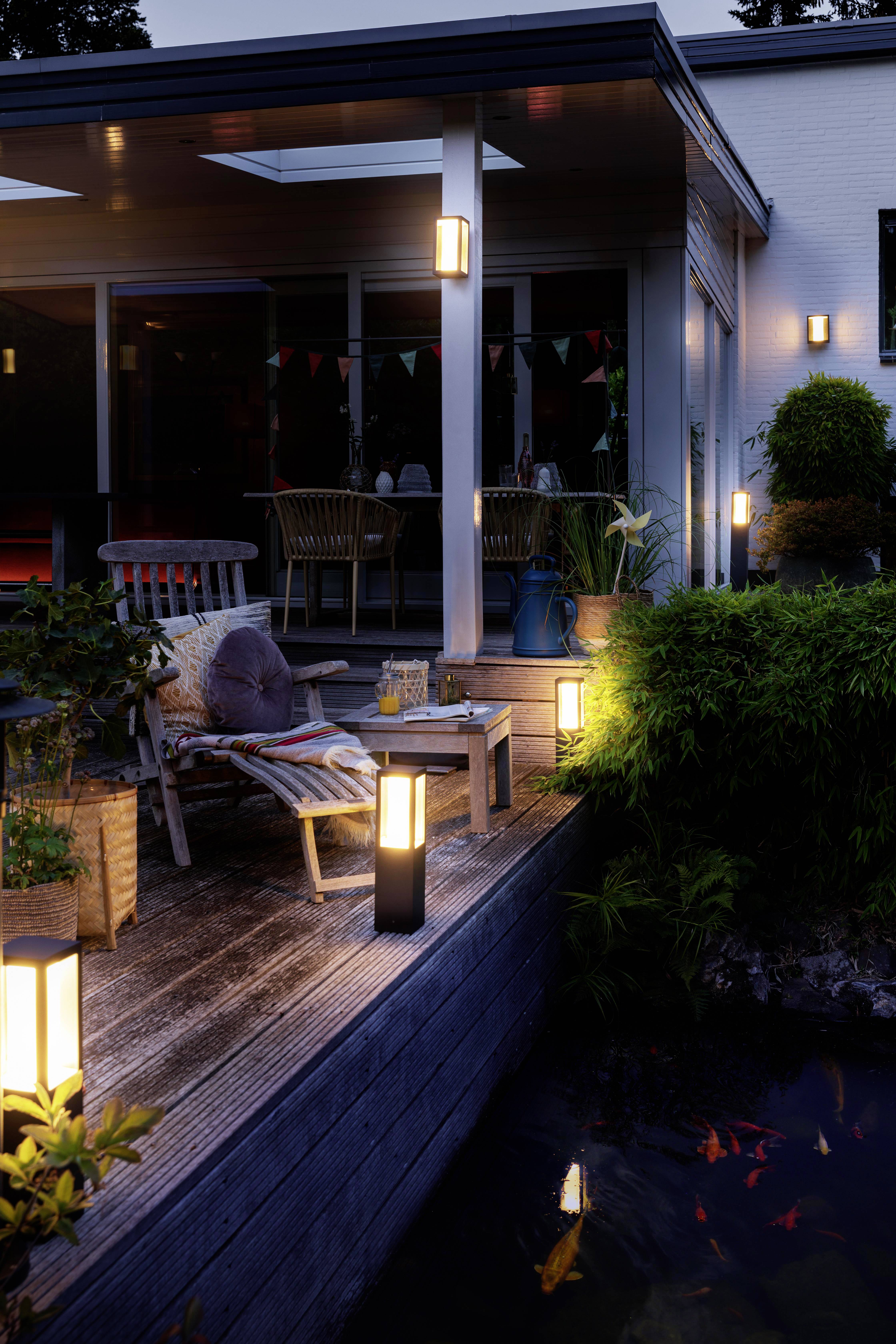 Eine abendliche Terrasse mit Beleuchtung. Loungemöbel und Tisch stehen auf dem Holzdeck. Im Vordergrund ist ein Teich mit Fischen sichtbar.