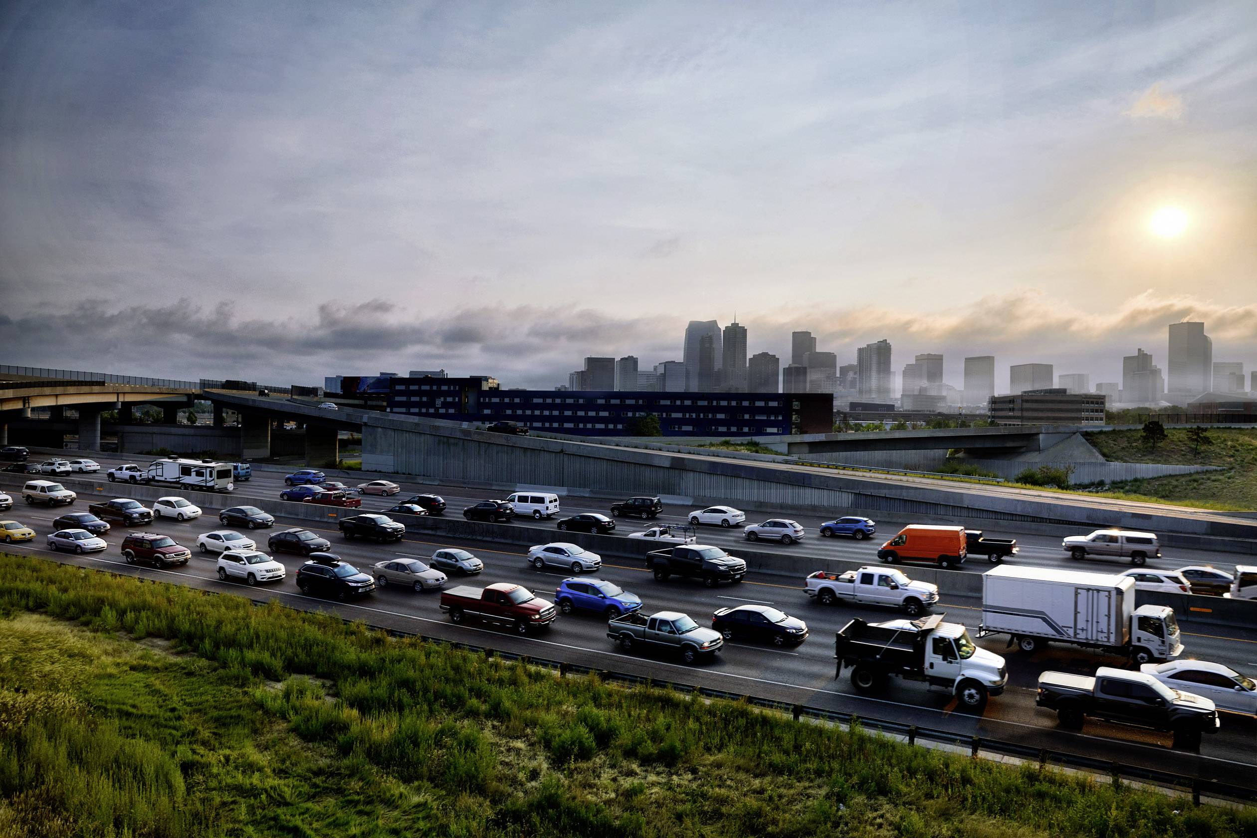 Dichter Verkehr auf einer Autobahn bei Sonnenuntergang, mit einer Stadt und Hochhäusern im Hintergrund. Hauptsächlich Autos und LKWs.