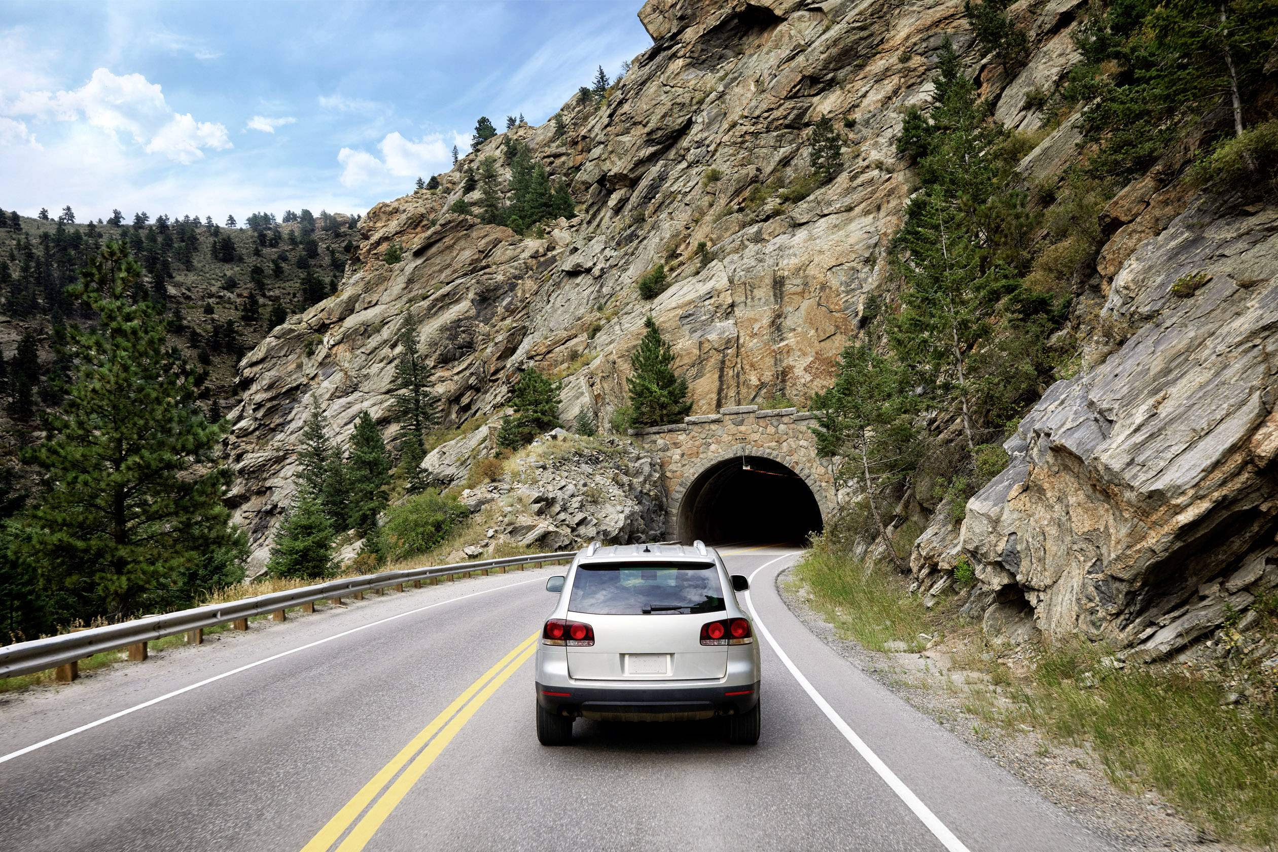 Ein Auto fährt auf einer Straße durch eine felsige Landschaft, mit einem Tunnel im Hintergrund, umgeben von Bäumen und blauen Himmel.