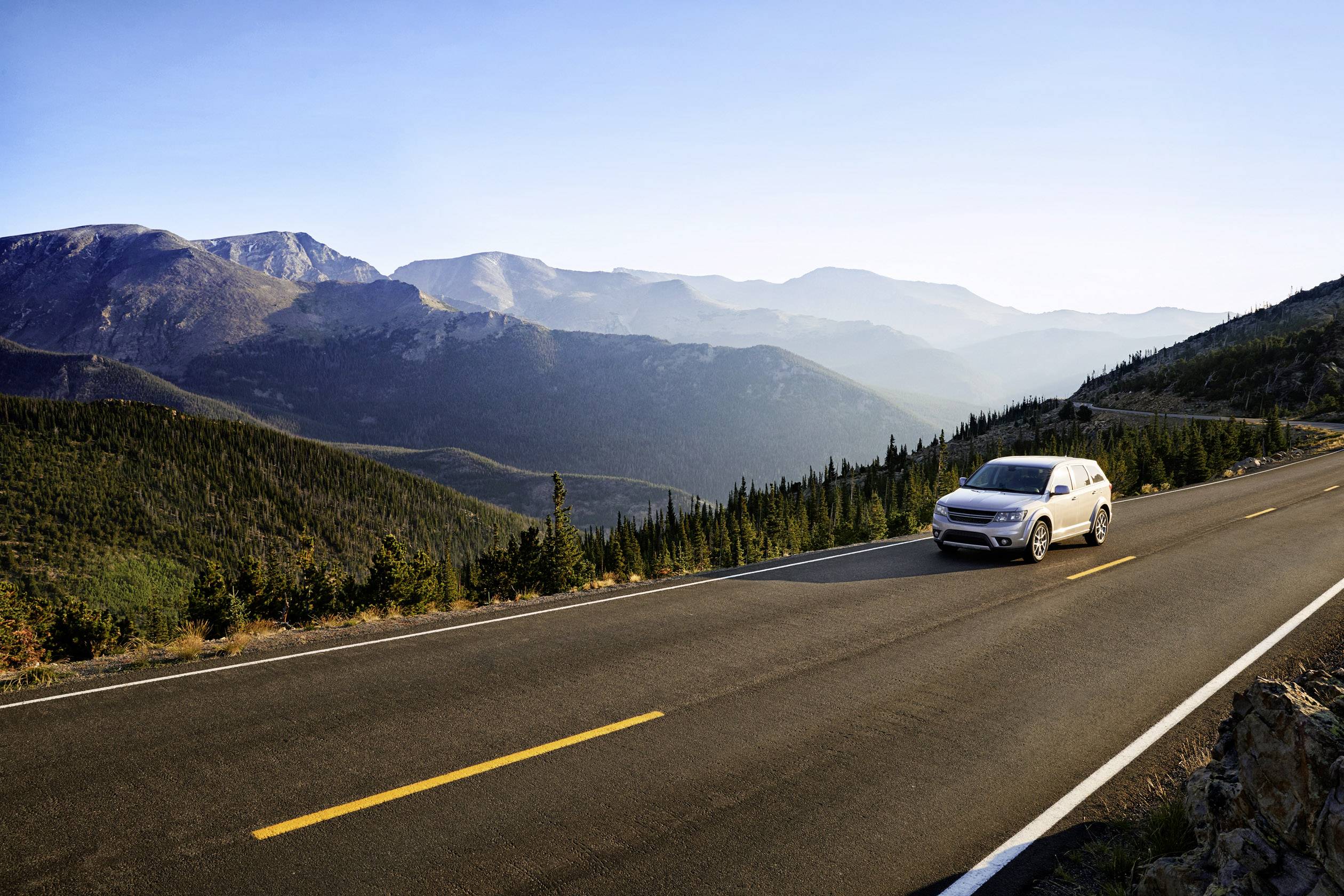 Ein silbernes Auto fährt auf einer kurvenreichen Straße durch eine bergige Landschaft mit bewaldeten Hügeln und blauem Himmel im Hintergrund.