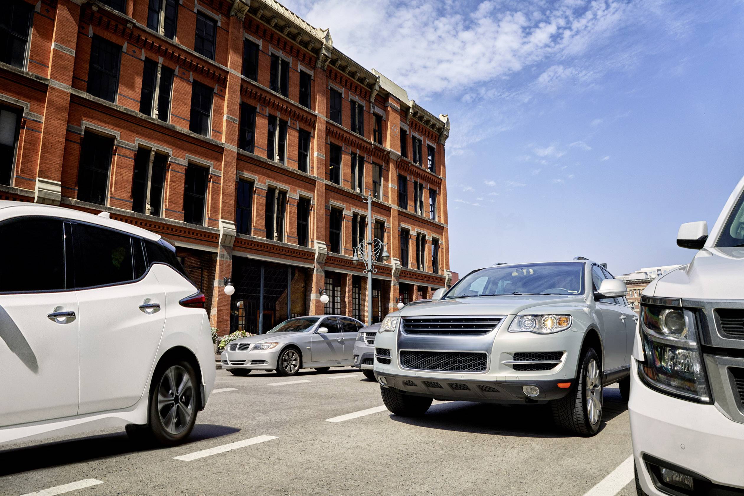 Mehrspurige Straße in einer Stadt, gesäumt von roten Backsteingebäuden und mehreren Pkw. Klarer Himmel im Hintergrund.