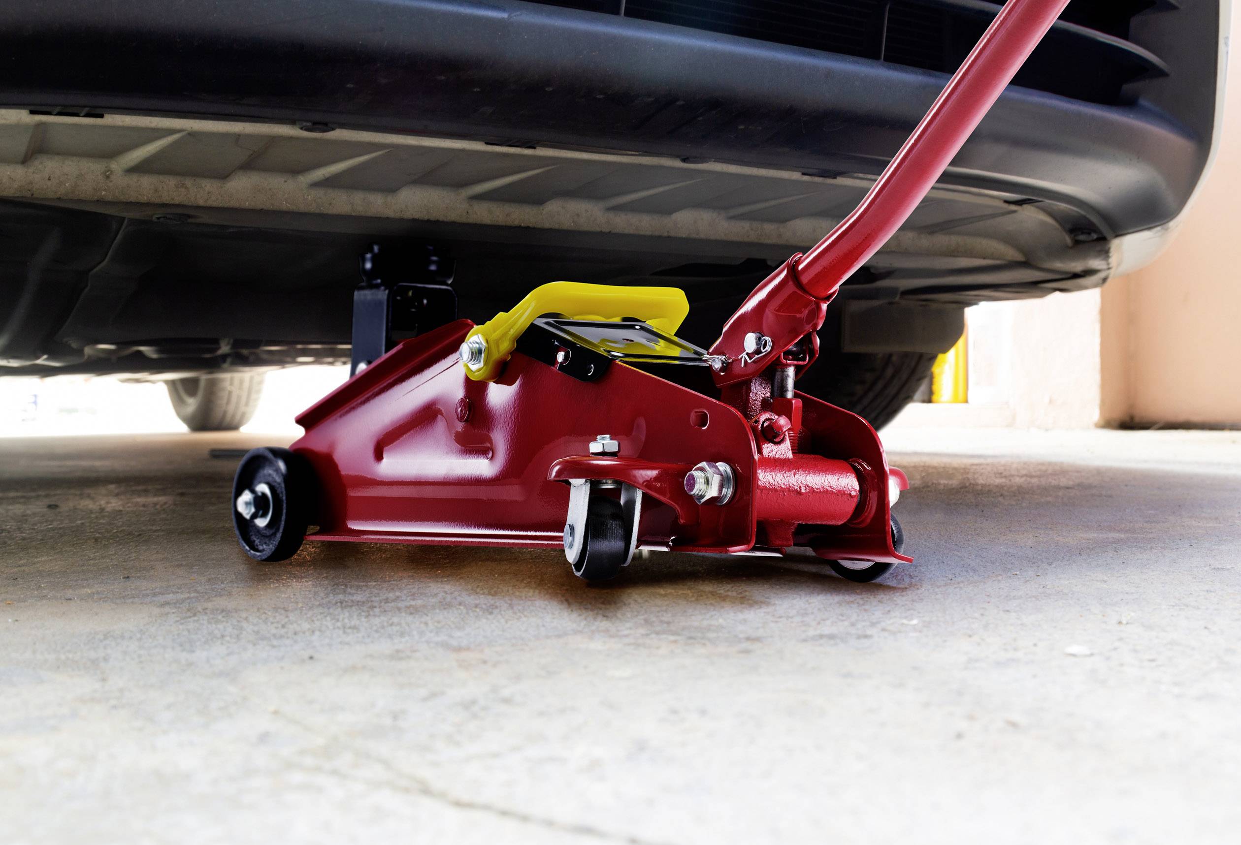 Ein roter Wagenheber hebt ein Auto in einer Garage an. Der Wagenheber befindet sich unter dem vorderen Teil des Fahrzeugs.