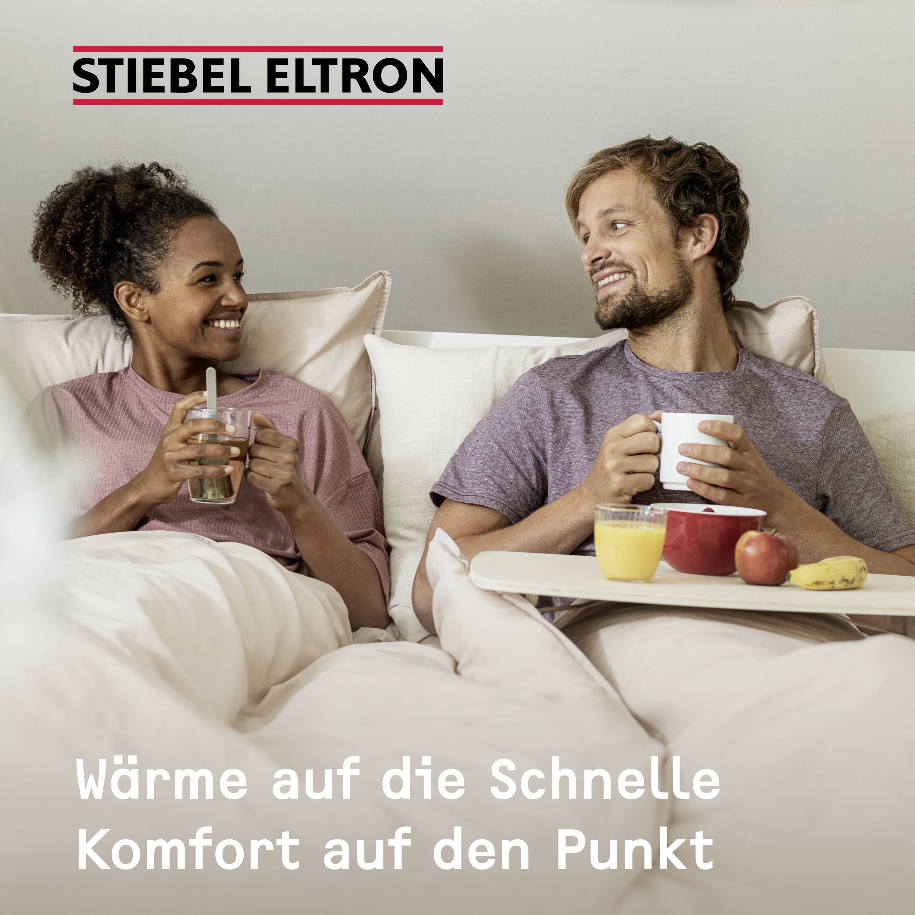 Ein lächelndes Paar sitzt auf einem Sofa und hält Getränke. Die Frau hat lockiges Haar, der Mann hat kurzes Haar und trägt einen Bart.