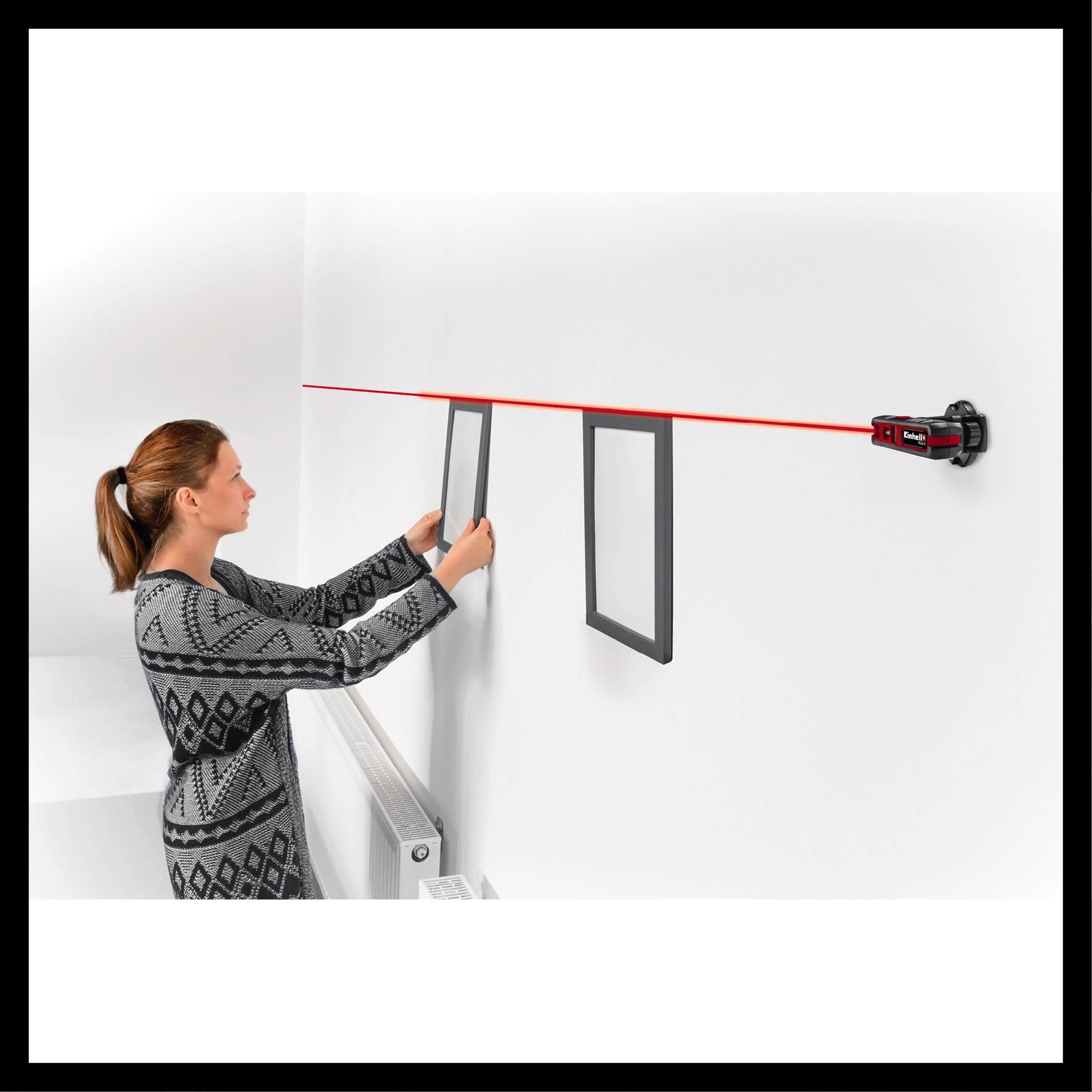 Eine Frau hängt mit Hilfe eines Lasernivelliergeräts zwei Bilder an einer weißen Wand auf.