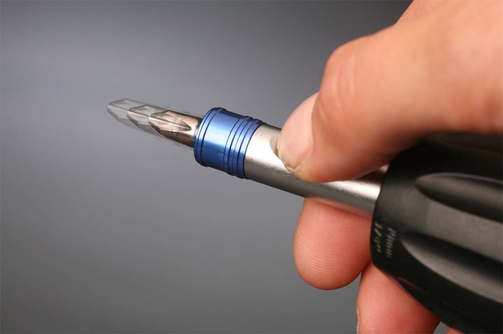 Eine Hand hält einen Schraubendreher mit einem blauen Ring und silberner Spitze, bereit zum Gebrauch gegen einen grauen Hintergrund.