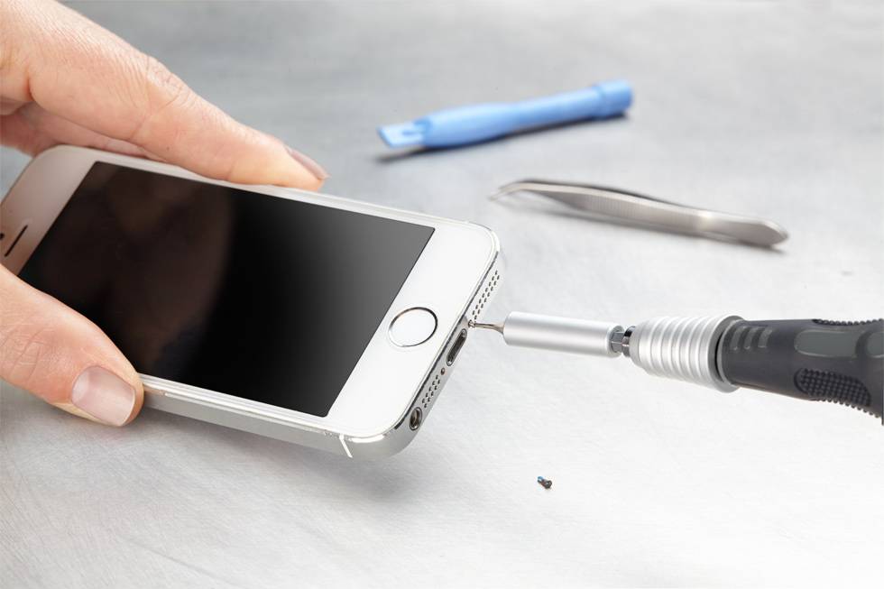 Eine Person repariert ein Smartphone mit einem Schraubendreher. Im Hintergrund liegen Werkzeuge wie eine Pinzette und ein Plastikhebel.