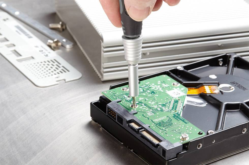 Eine Person repariert eine Festplatte mit einem Schraubenzieher. Im Hintergrund befinden sich weitere elektronische Komponenten.