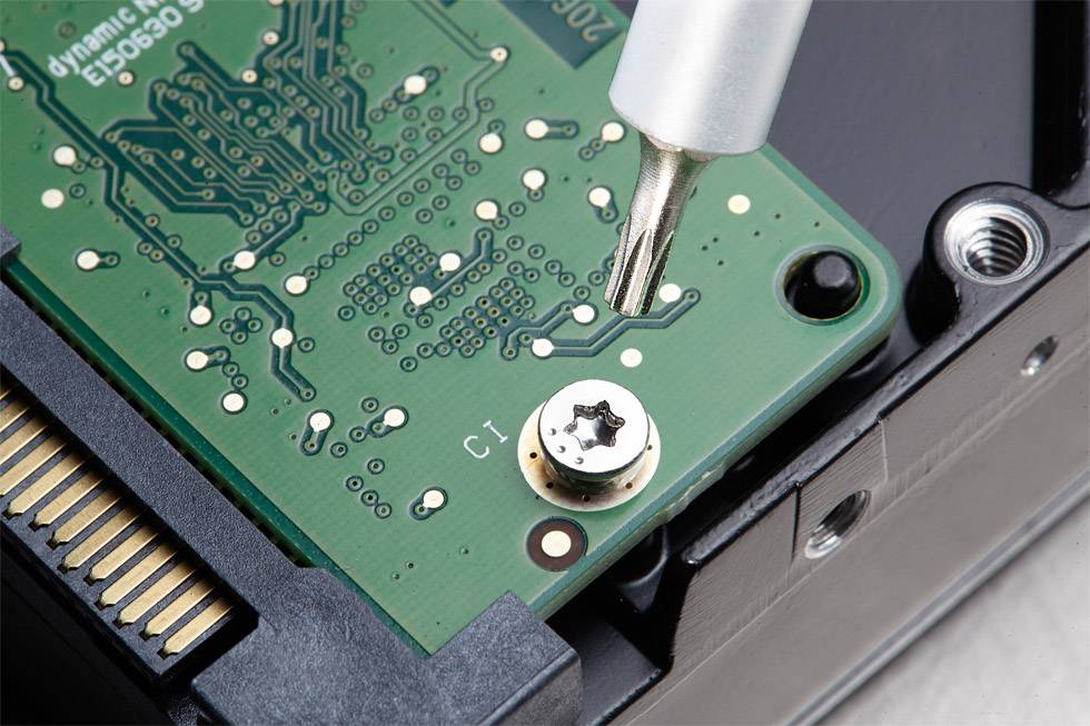 Nahaufnahme einer grünen Platine mit einem Metallwerkzeug, das eine Schraube anzieht. Links ist ein Teil eines elektronischen Anschlusses zu sehen.