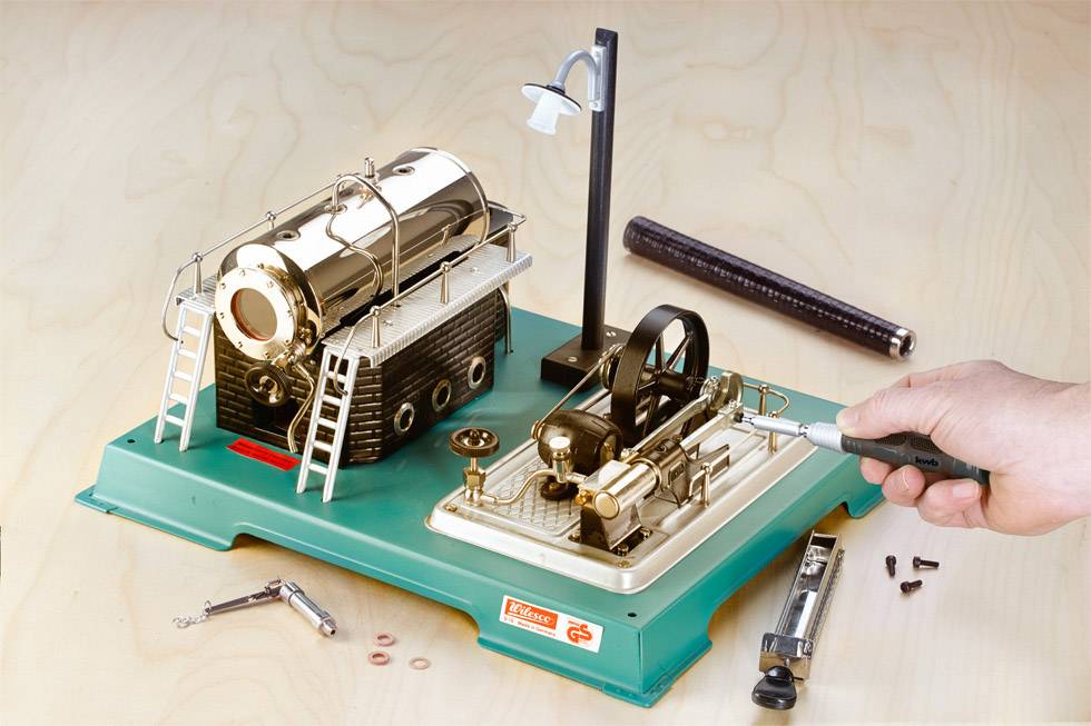 Spielzeug-Dampfmaschine-Set auf grünem Sockel, mit einem glänzenden Kessel und einem Kolben. Hand betätigt Hebel zur Bewegung.