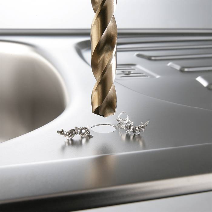 Bohrer durchdringt Edelstahlspüle; umgebende Metallspäne sichtbar.