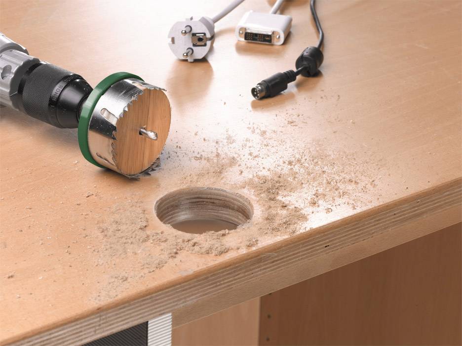 Ein Schreibtisch mit einem frisch gebohrten Loch zeigt Bohrwerkzeug und Holzspäne auf der Oberfläche. Kabel sind im Hintergrund sichtbar.