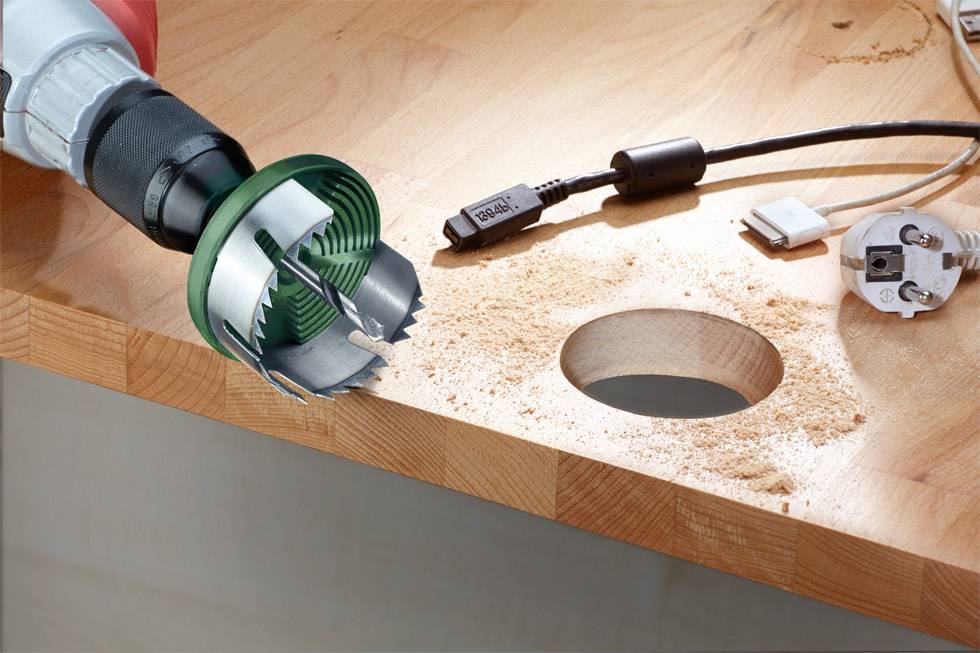 Bohrer mit Lochsägeaufsatz bohrt ein rundes Loch in eine Holzoberfläche, Holzspäne um das Loch. Ein Kabel und Stecker sind daneben.