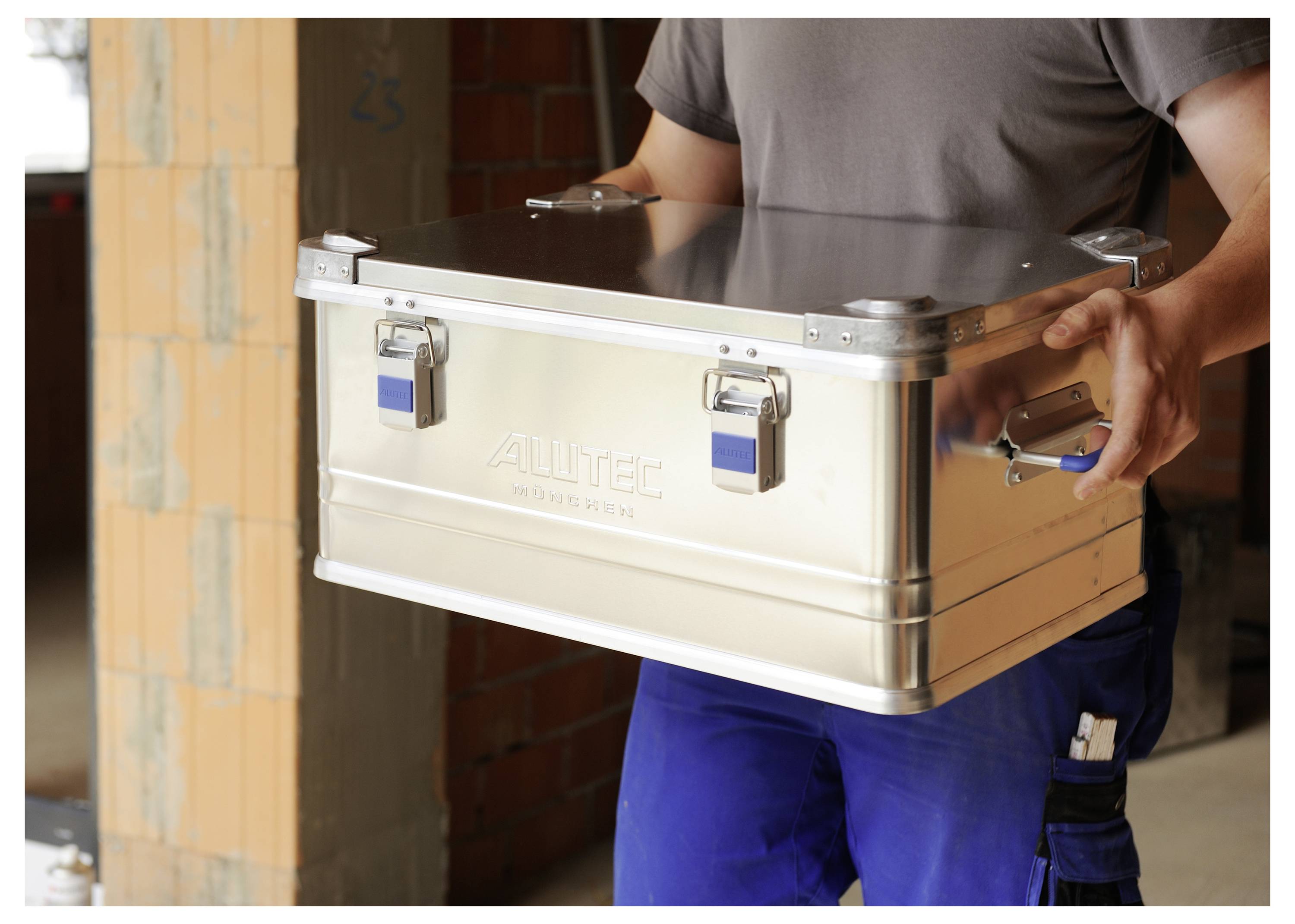 Eine Person, die eine Metalllagerkiste mit Seitengriffen hält, steht in einem teilweise errichteten Gebäude mit freiliegenden Ziegelwänden.