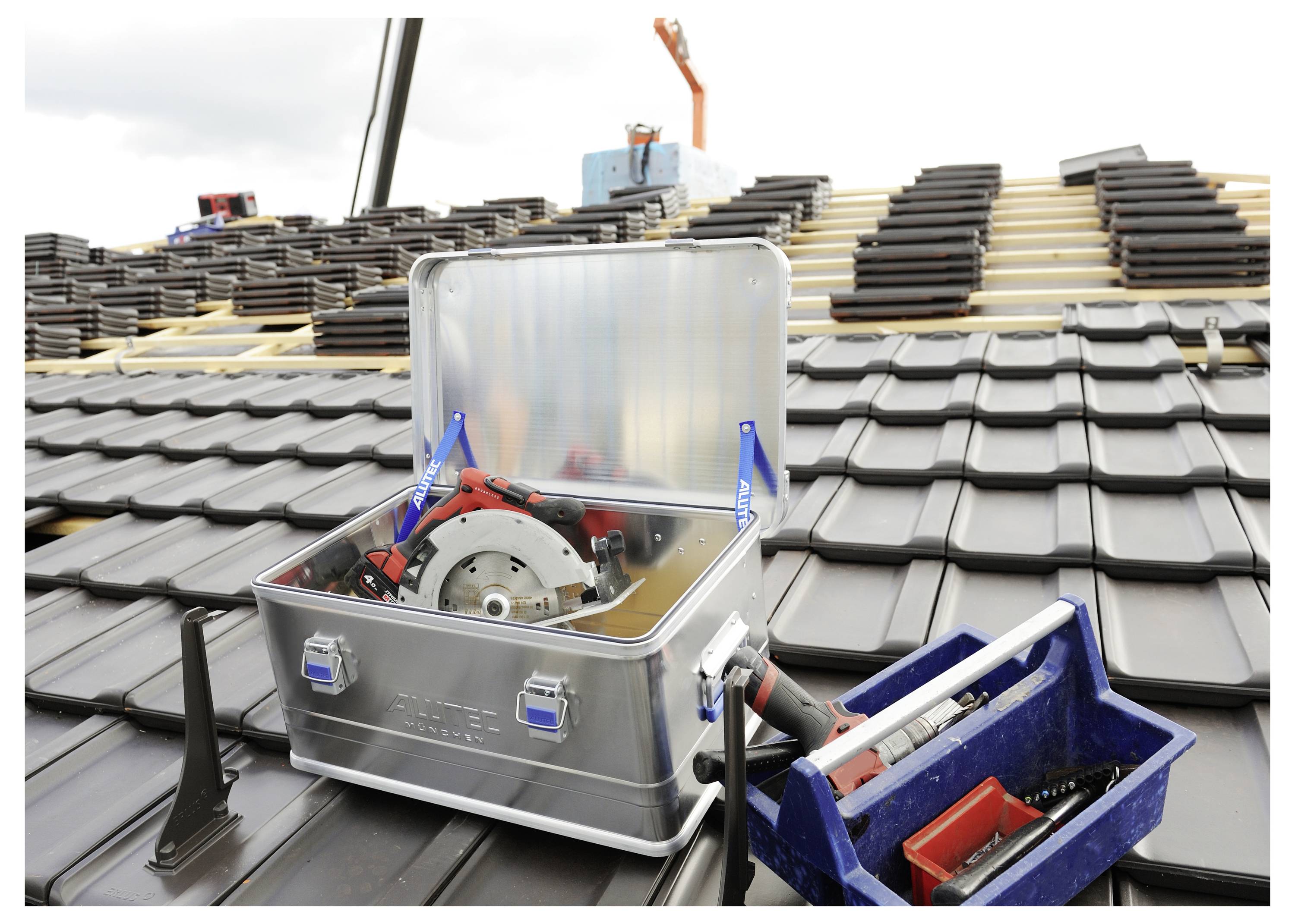 Eine offene Metallwerkzeugkiste mit einer Kreissäge und roten Griffen steht auf einem Ziegeldach, daneben ein blauer Behälter mit Werkzeugen.