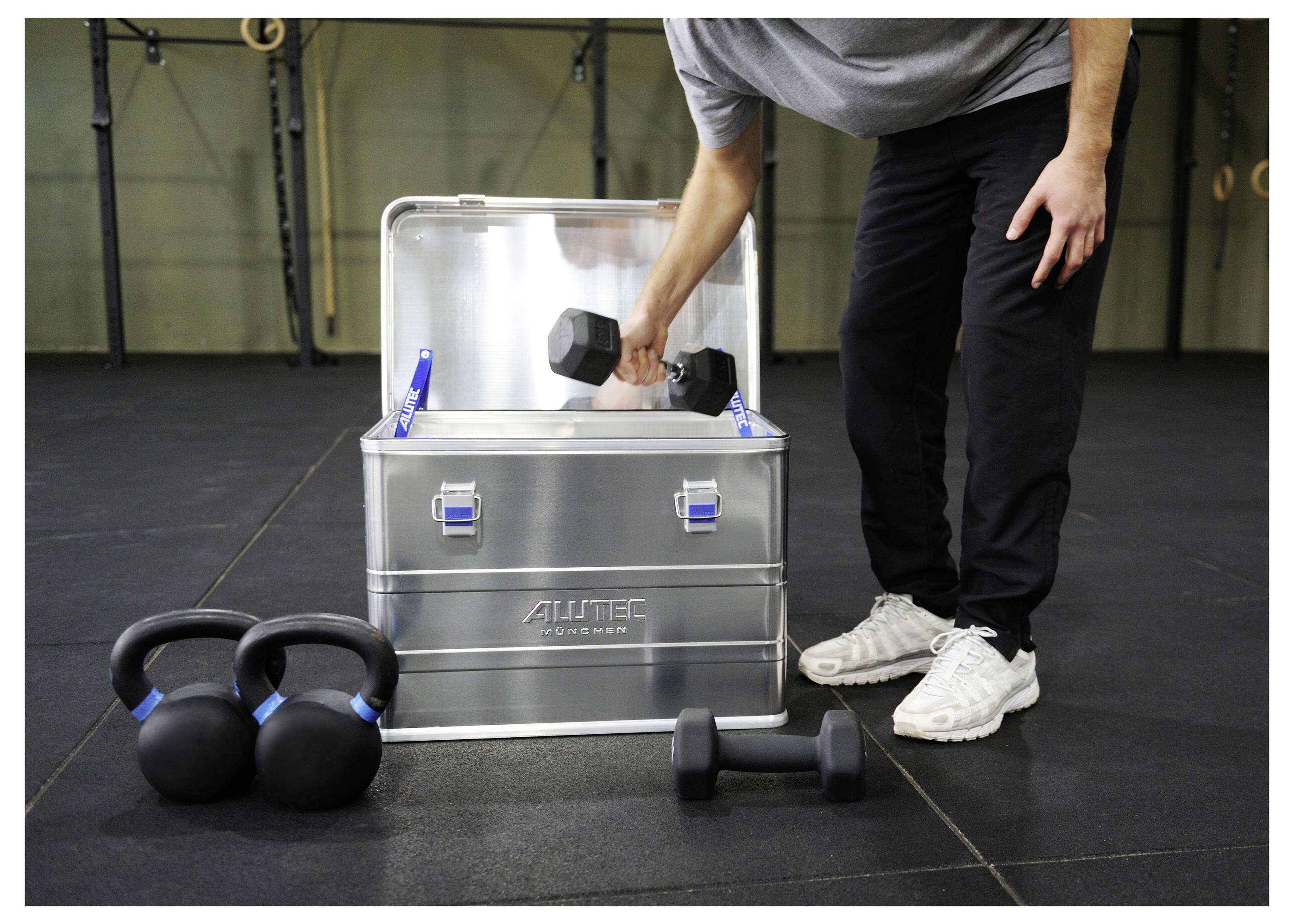 Eine Person legt eine Hantel in eine Metalllagerkiste in einer Sporthallumgebung, während Kettlebells und eine weitere Hantel in der Nähe auf dem Boden liegen.