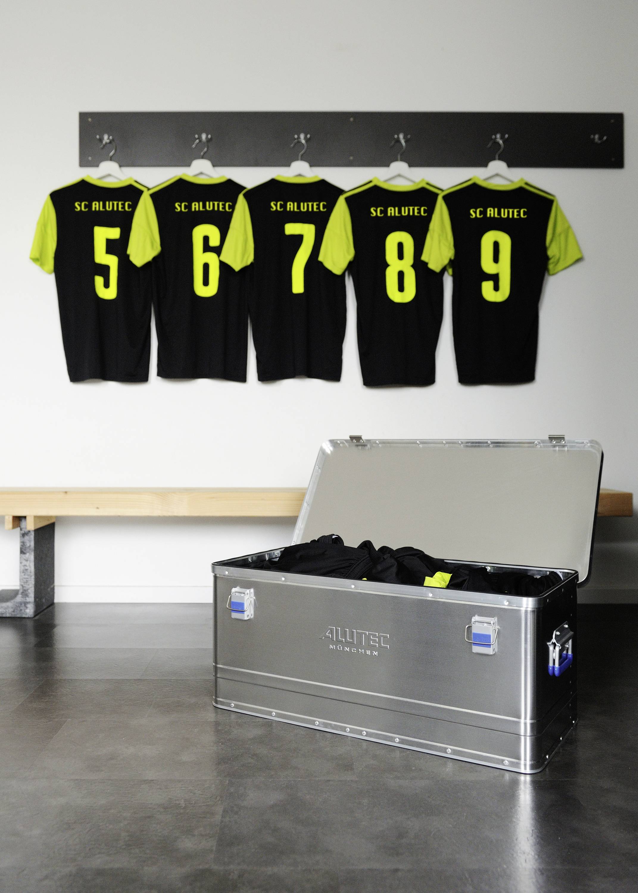 Fünf schwarze Sporttrikots mit gelben Nummern (5-9) hängen an der Wand, daneben eine geöffnete Metallkiste mit weiteren Trikots.