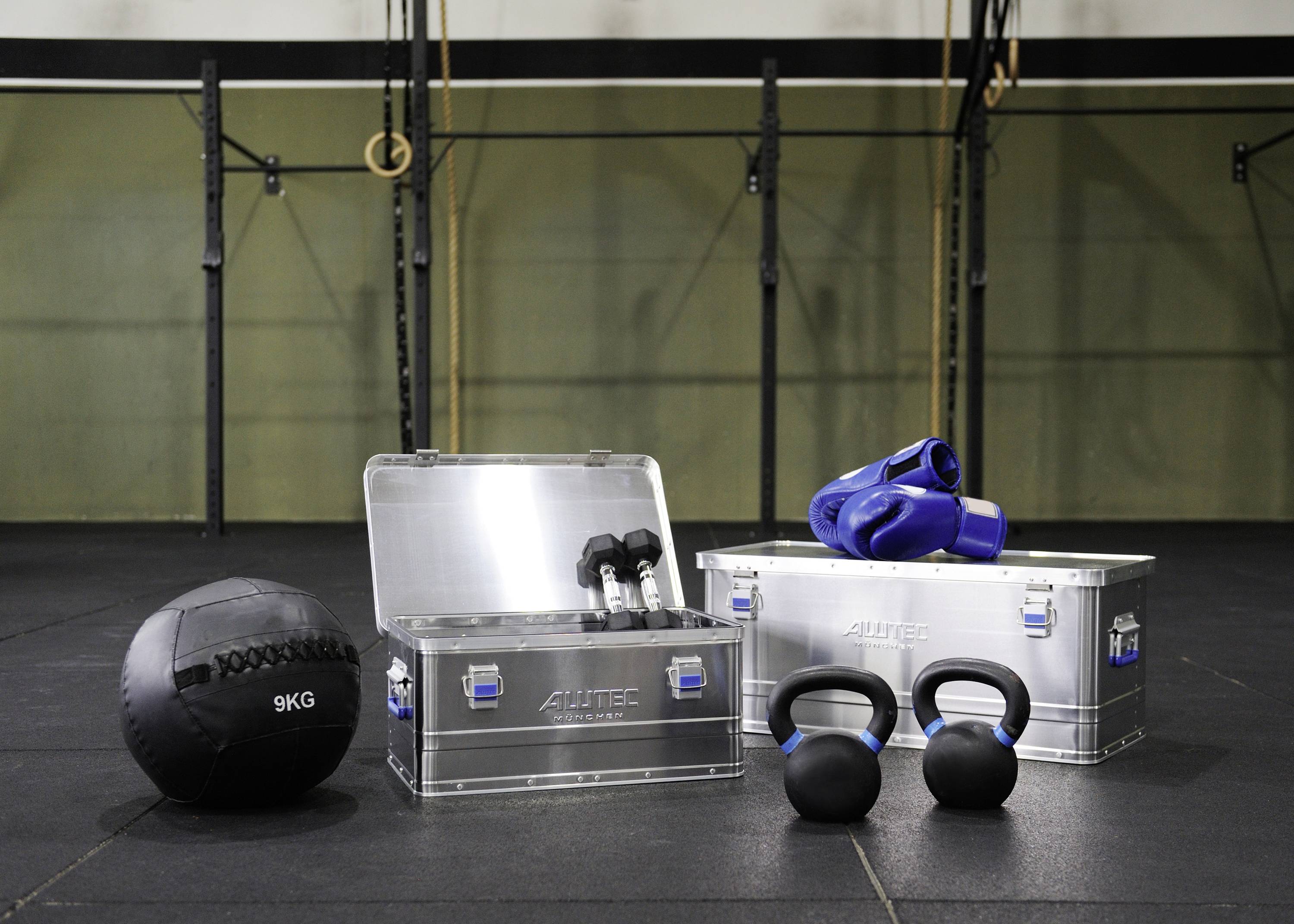 Fitnessstudio mit CrossFit-Ausrüstung: Medizinball, Kettlebells, Aluminiumboxen und Sporthandschuhe auf Boden vor Ringen und Klimmzugstangen.