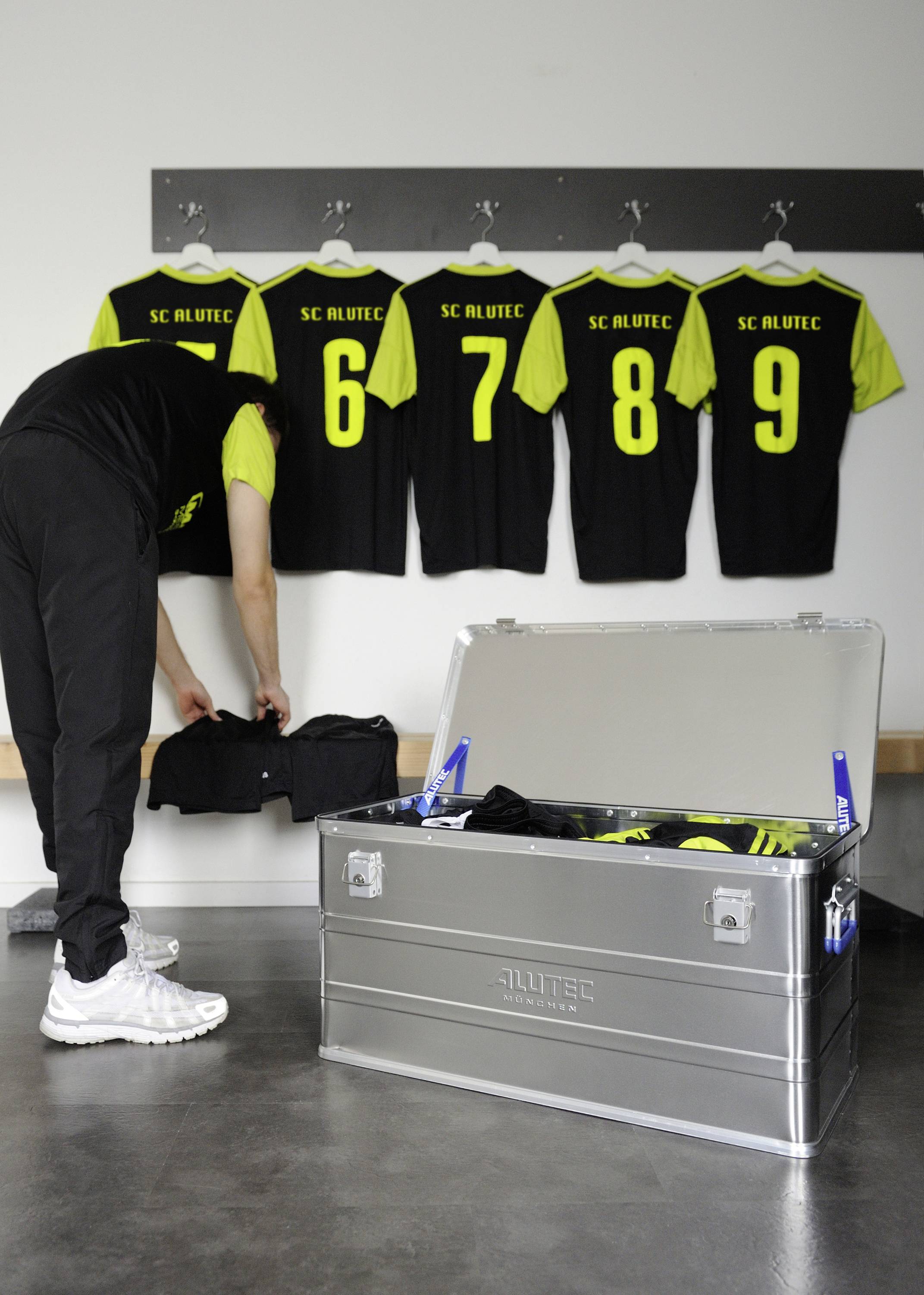 Eine Person holt Fußballtrikots aus einer silbernen Kiste in einer Umkleidekabine; im Hintergrund hängen nummerierte Trikots an Haken.