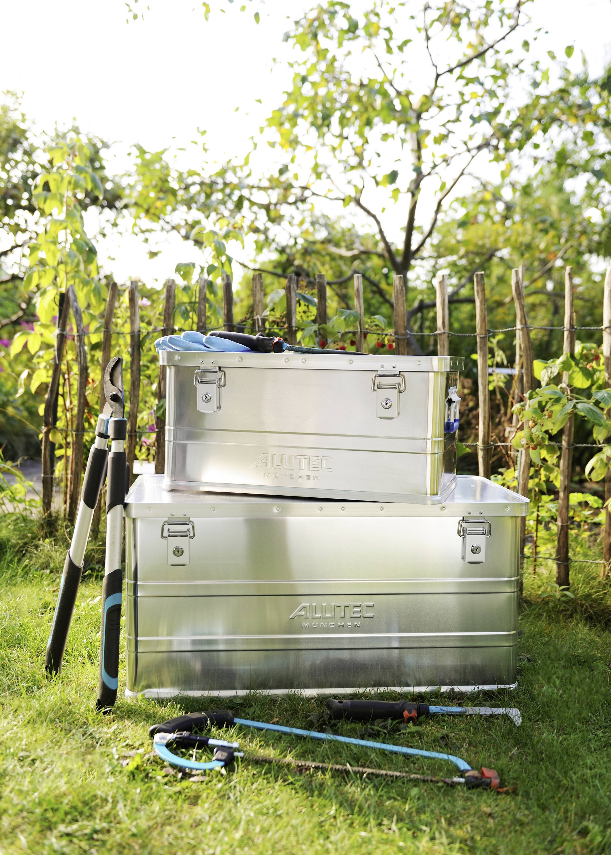 Zwei Metallboxen und Gartenwerkzeuge stehen auf dem Rasen vor einem Holzzaun. Perfekt für Gartenlagerung und Organisation.