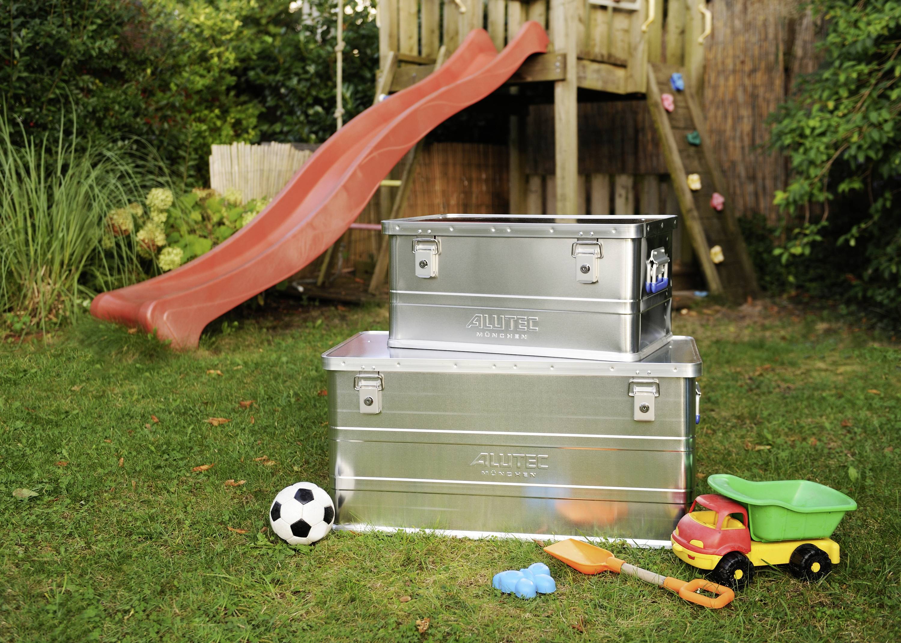 Zwei Metallkisten auf einer Wiese vor einer roten Rutsche, umgeben von Kinderspielzeug wie einem Fußball und einem Spielzeugauto.