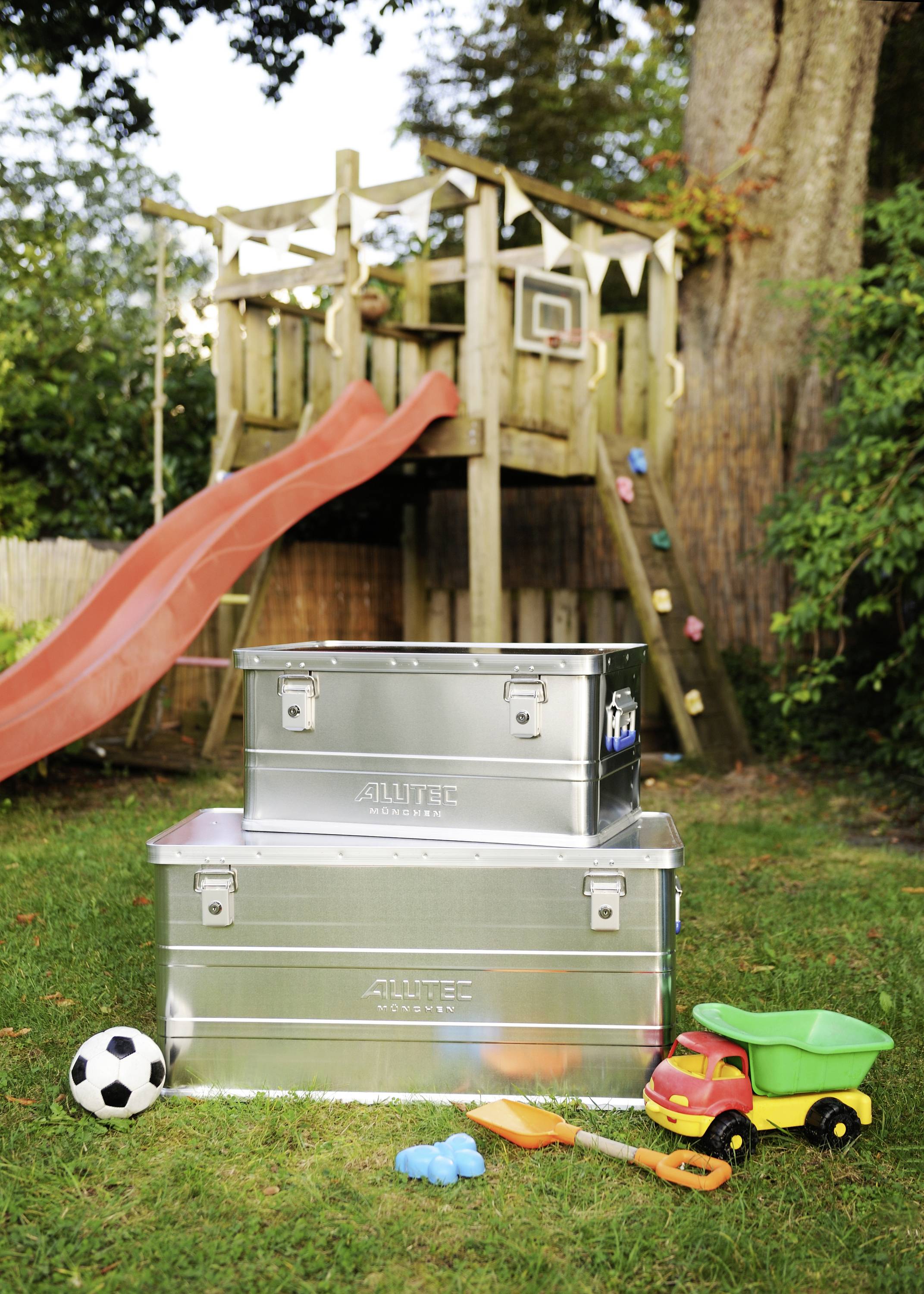 Zwei silberne Kisten stehen im Garten vor einem Klettergerüst mit Rutsche; daneben ein Fußball, Spielzeugauto und Schaufeln.