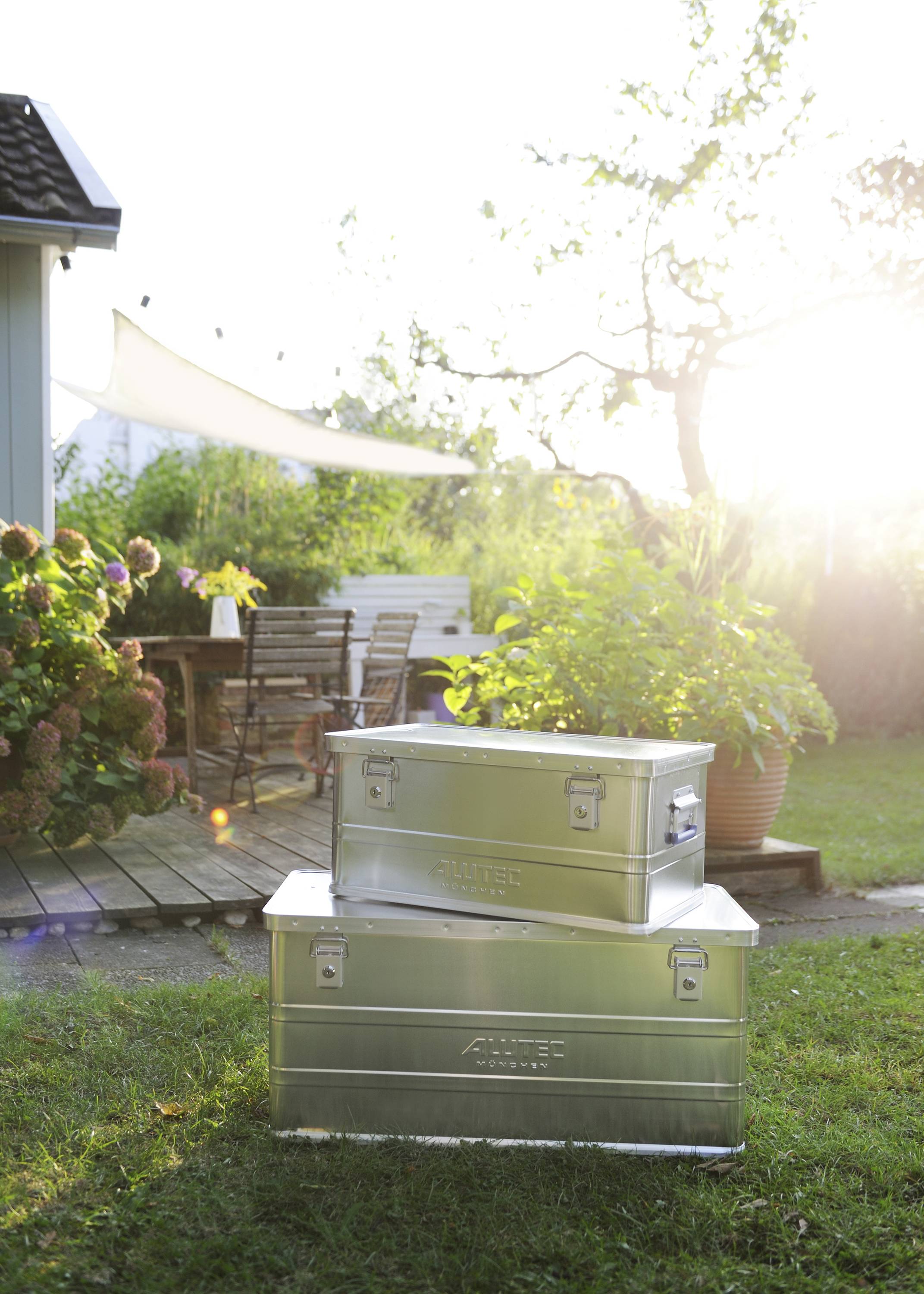 Zwei silberne Metallboxen stehen gestapelt auf grünem Rasen vor einer Terrasse mit Holztisch und Stühlen im Sonnenlicht.