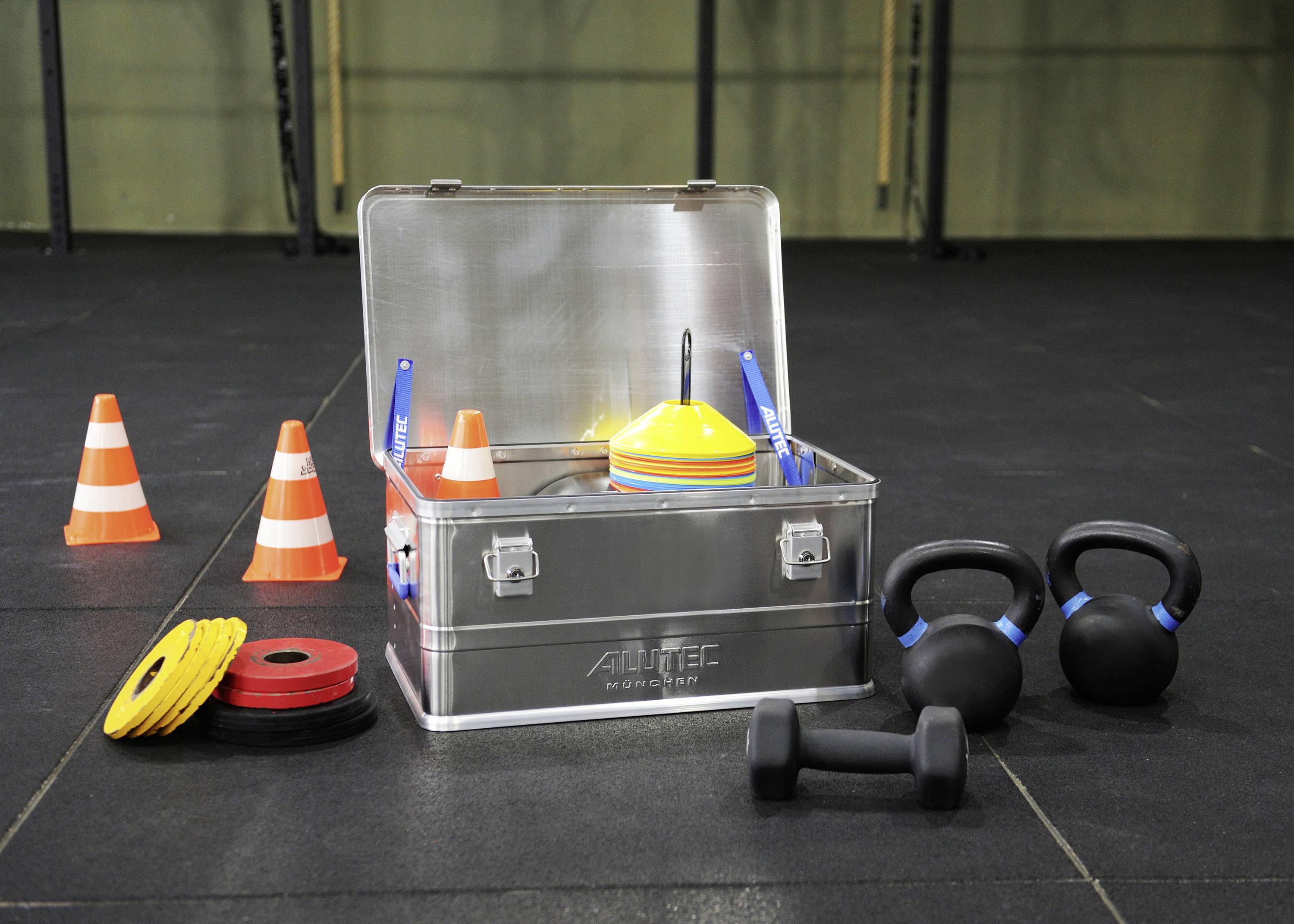 Fitness-Ausrüstung: Metallbox mit Hanteln, Kettlebells, Hütchen und Gewichtsplatten in einer Turnhalle, bereit für ein Workout.