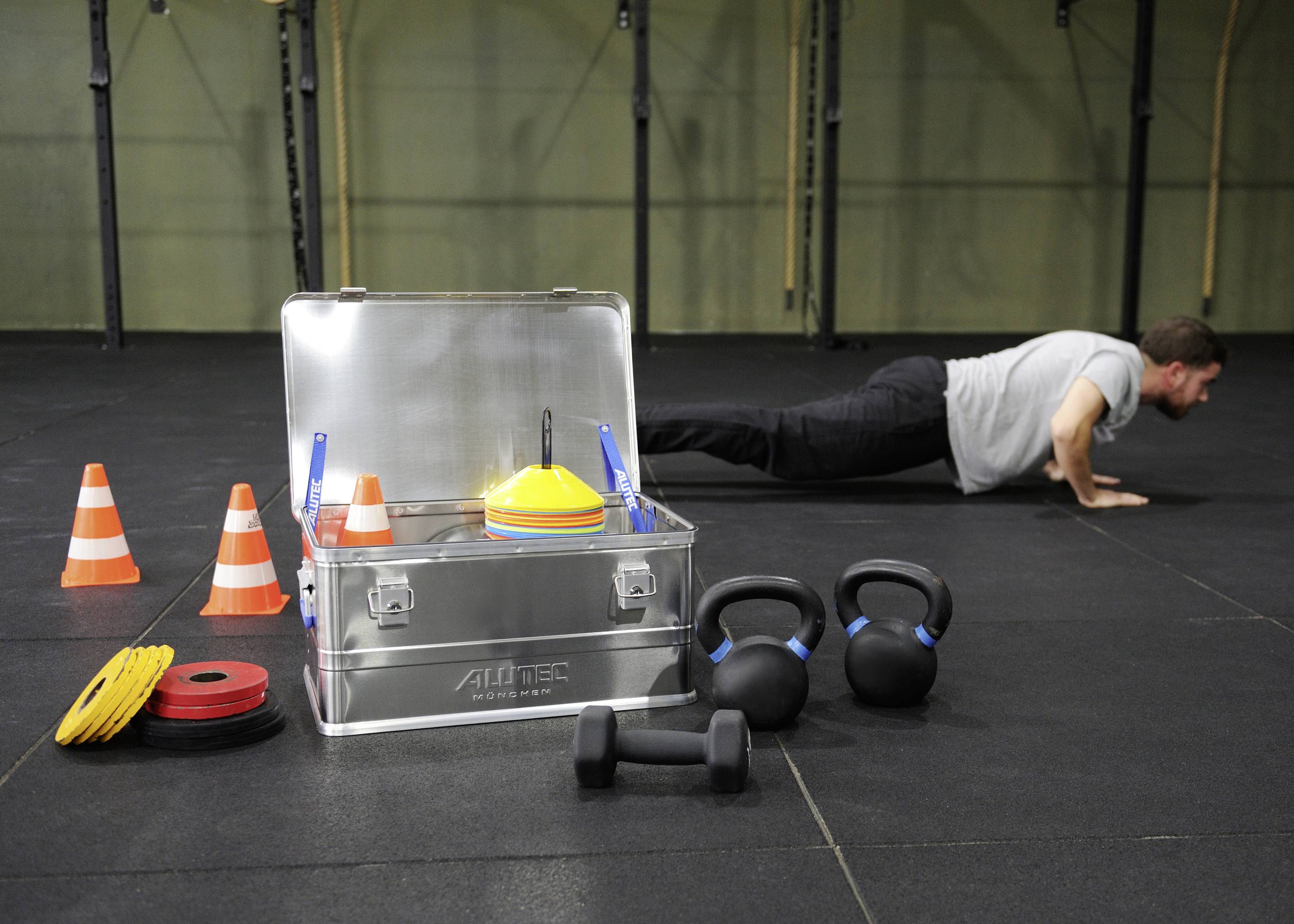 Ein Mann macht Liegestütze in einem Fitnessstudio. Neben ihm stehen Kettlebells, bunte Kegel und eine offene Metallbox mit Sportgeräten.