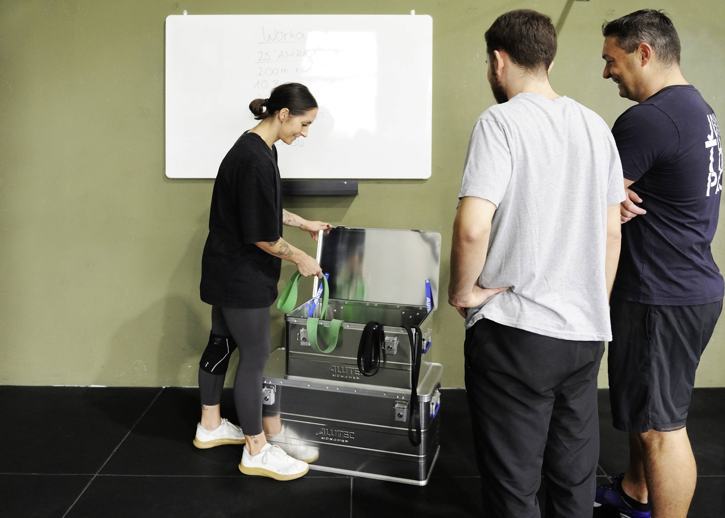 Eine Person zeigt zwei weiteren Personen etwas in einer Kiste in einem Fitnessstudio. Ein Whiteboard hängt an der Wand im Hintergrund.