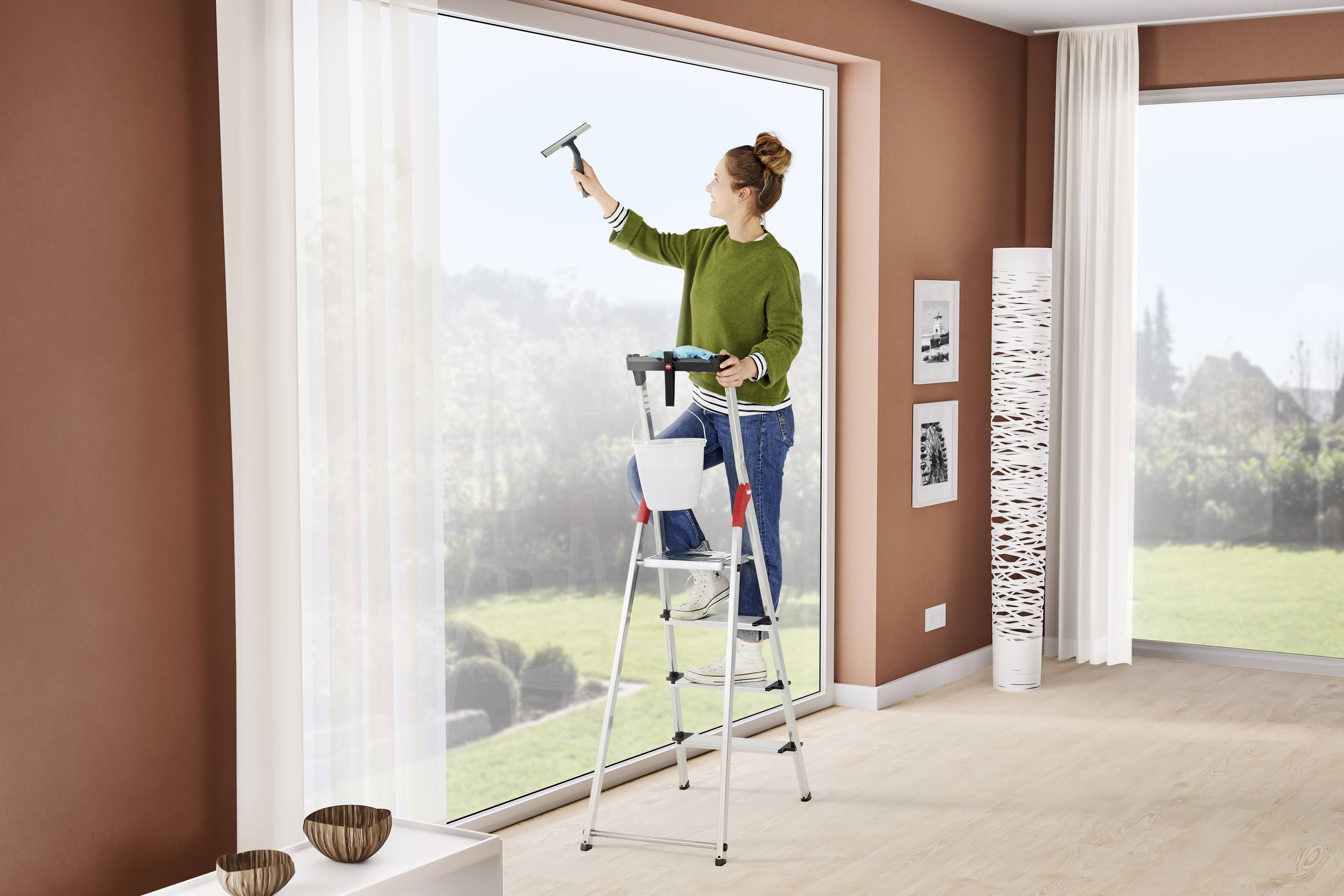 Eine Person steht auf einer Leiter und reinigt ein großes Fenster in einem modern eingerichteten Raum. Helle Wände und große Fenster bieten viel Tageslicht.