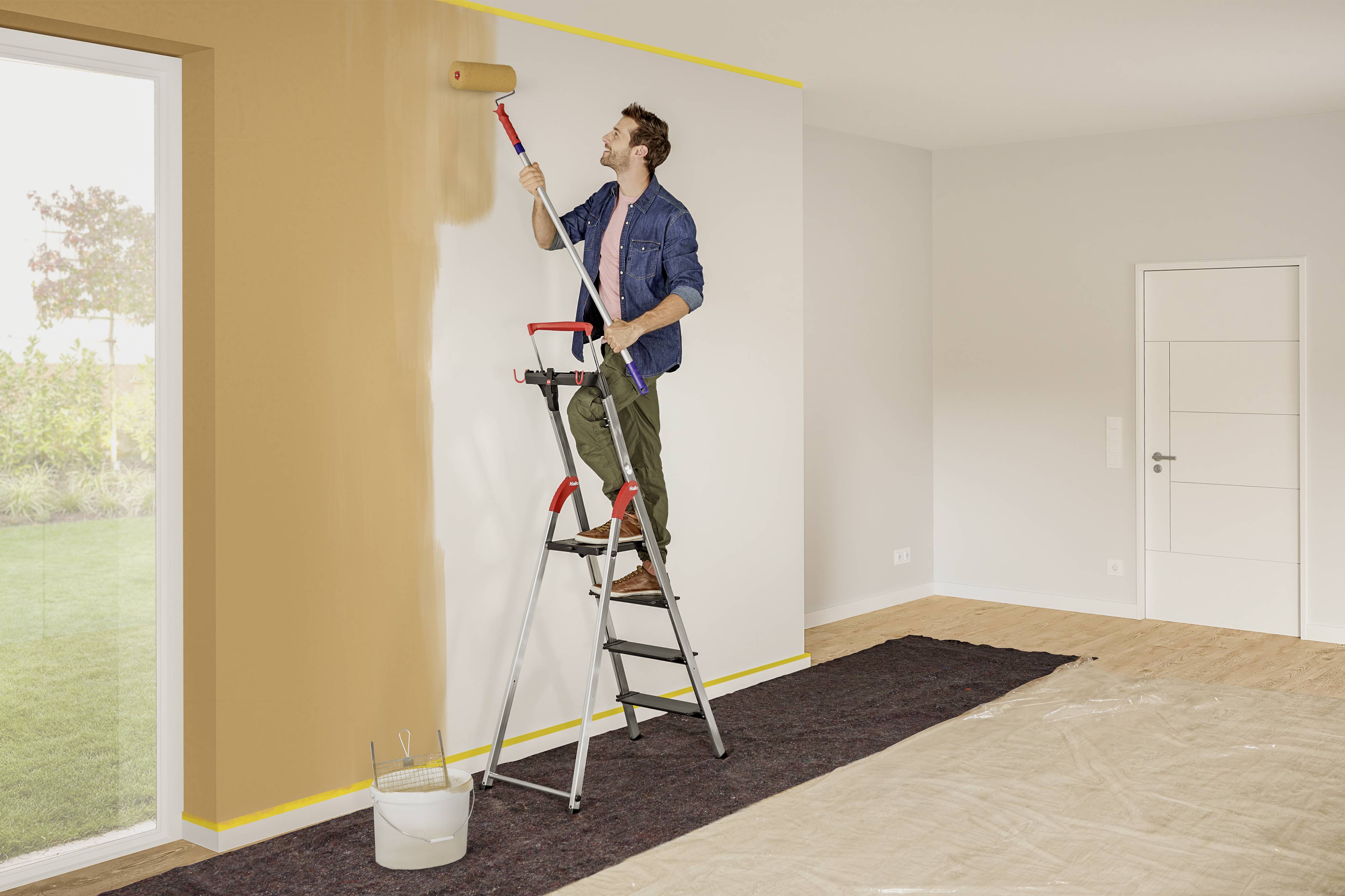 Ein Mann steht auf einer Leiter und streicht eine Wand in einem Raum von weiß zu orange. Neben ihm stehen ein Eimer und eine Farbrolle.