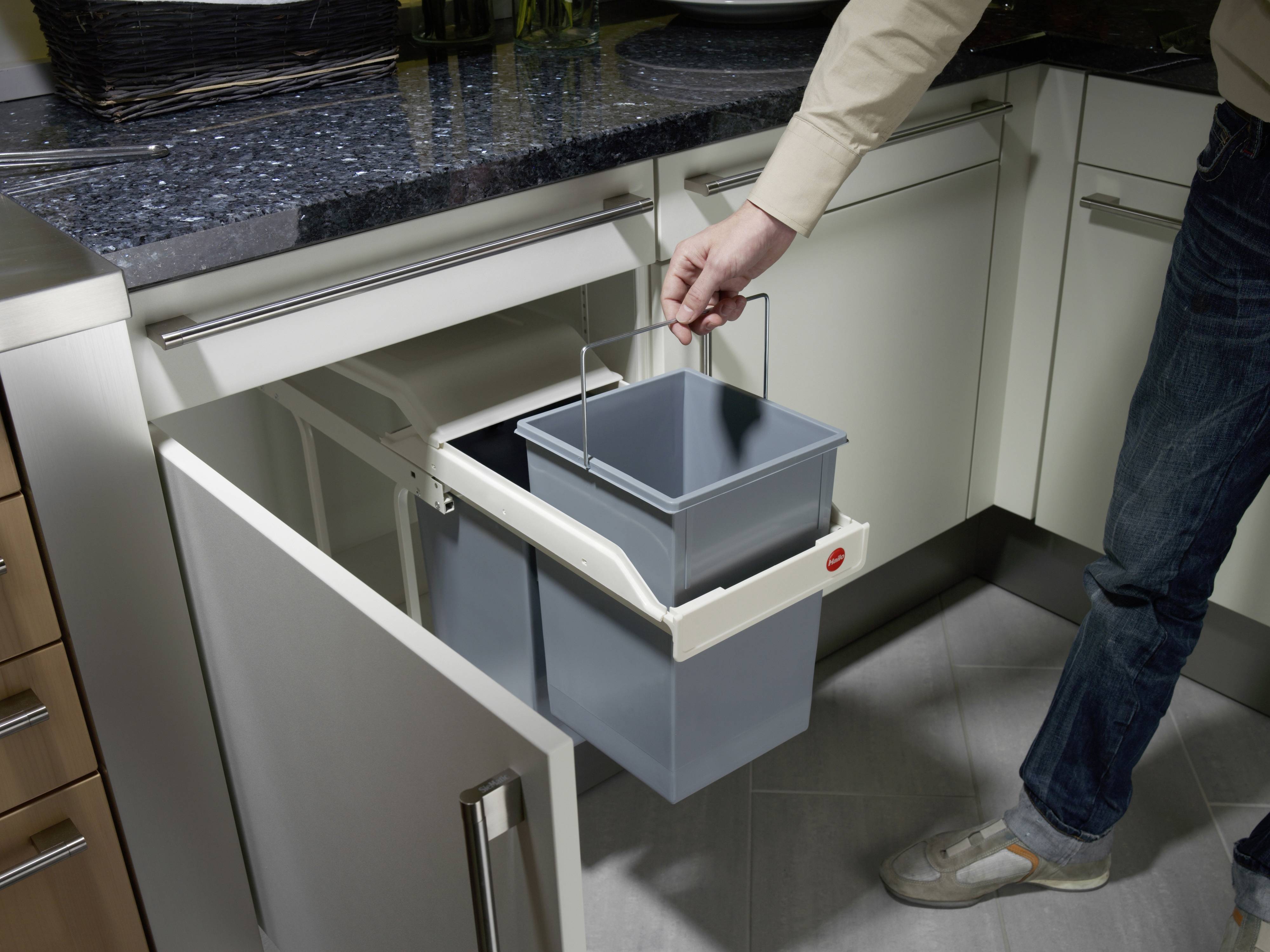 Eine Person öffnet einen Schrank unter der Spüle und hebt einen herausnehmbaren Mülleimer aus einem Abfalltrennsystem heraus.