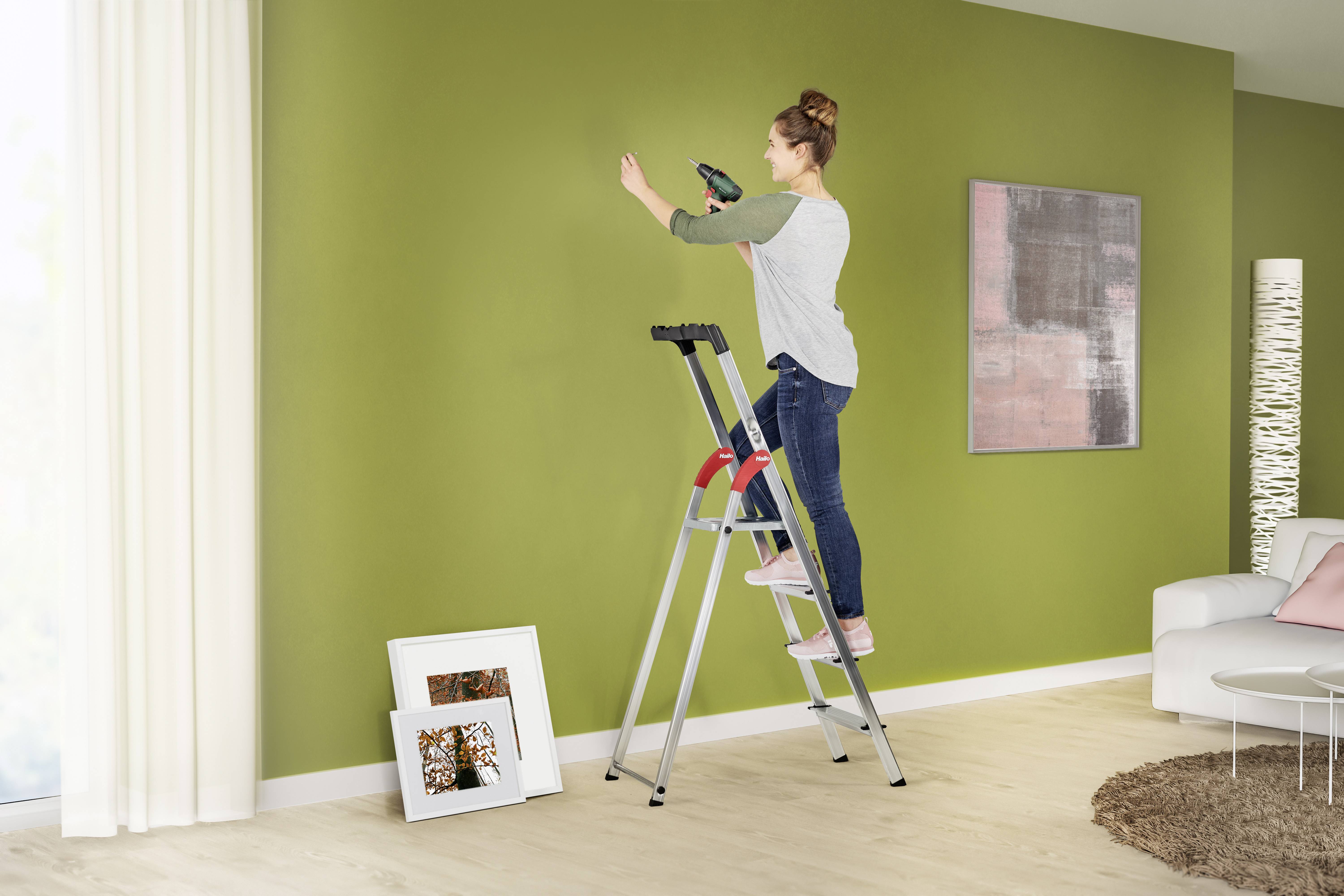 Eine Frau steht auf einer Leiter und bohrt ein Loch in eine grüne Wand. Unten sind gerahmte Bilder und im Hintergrund ein Sofa sichtbar.