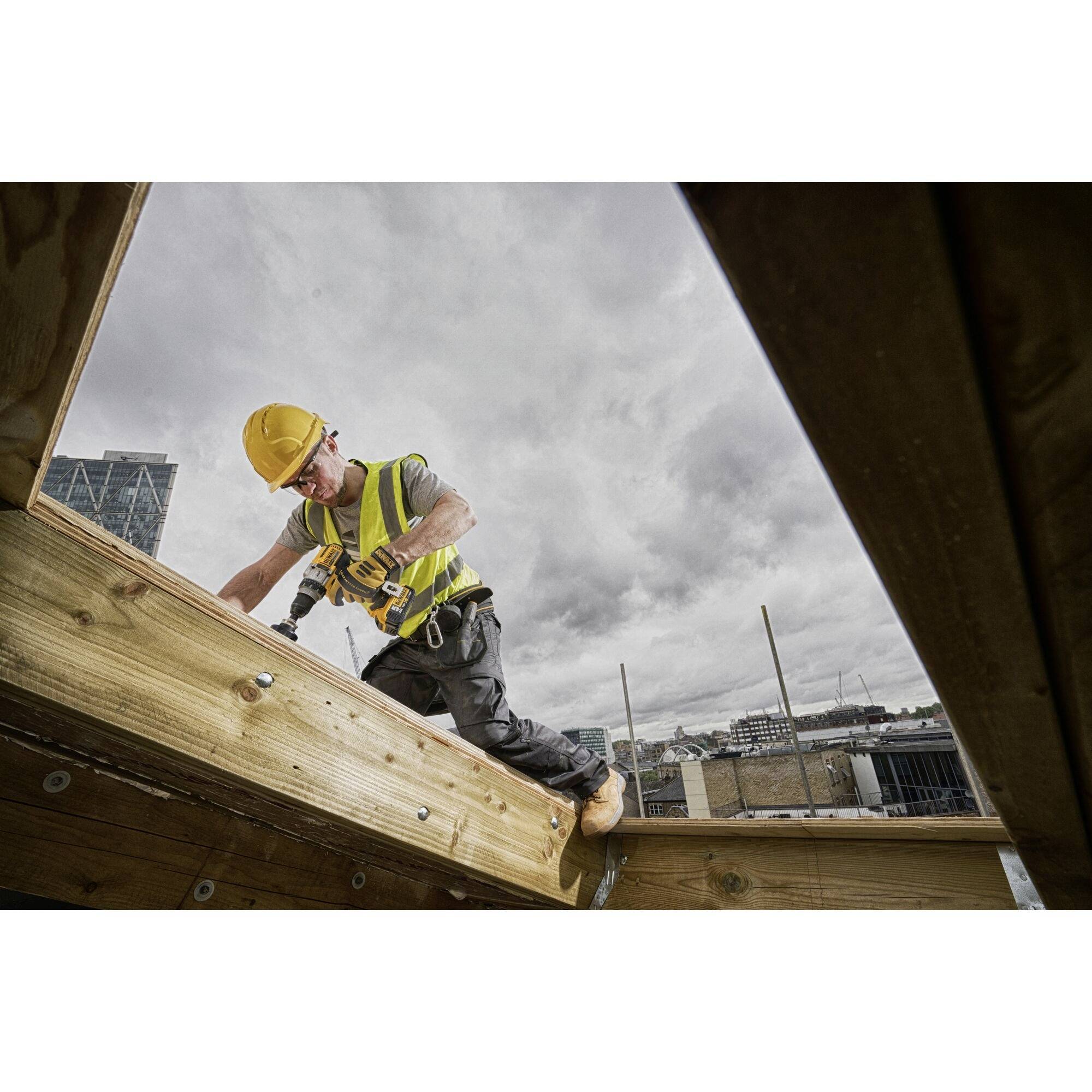 Ein Bauarbeiter mit Schutzhelm und Warnweste befestigt Holzplanken mit einer Bohrmaschine auf einer Baustelle unter bewölktem Himmel.