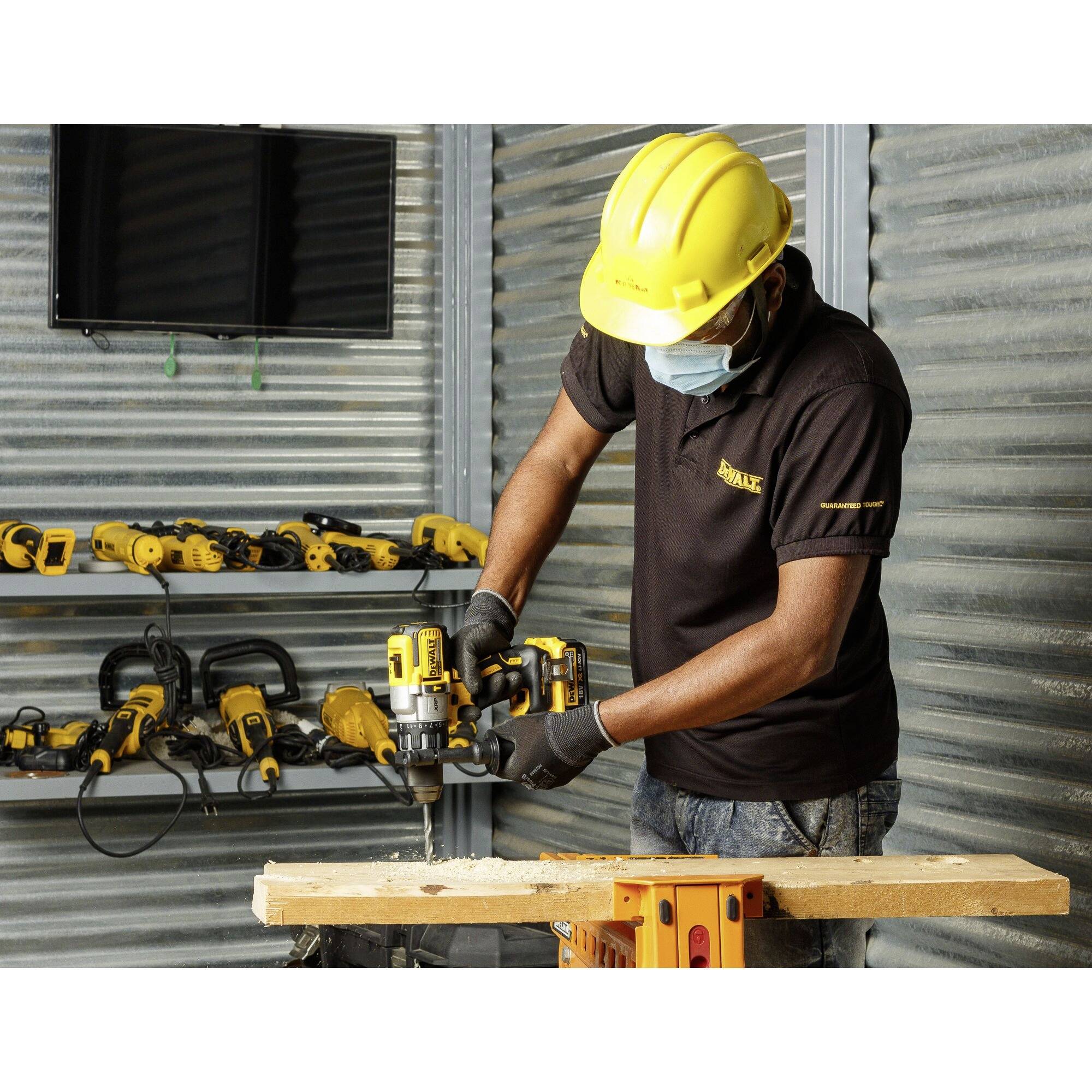 Ein Handwerker mit Schutzhelm und Maske arbeitet mit einem Elektrowerkzeug an einem Holzstück, im Hintergrund mehrere Werkzeuge an der Wand.
