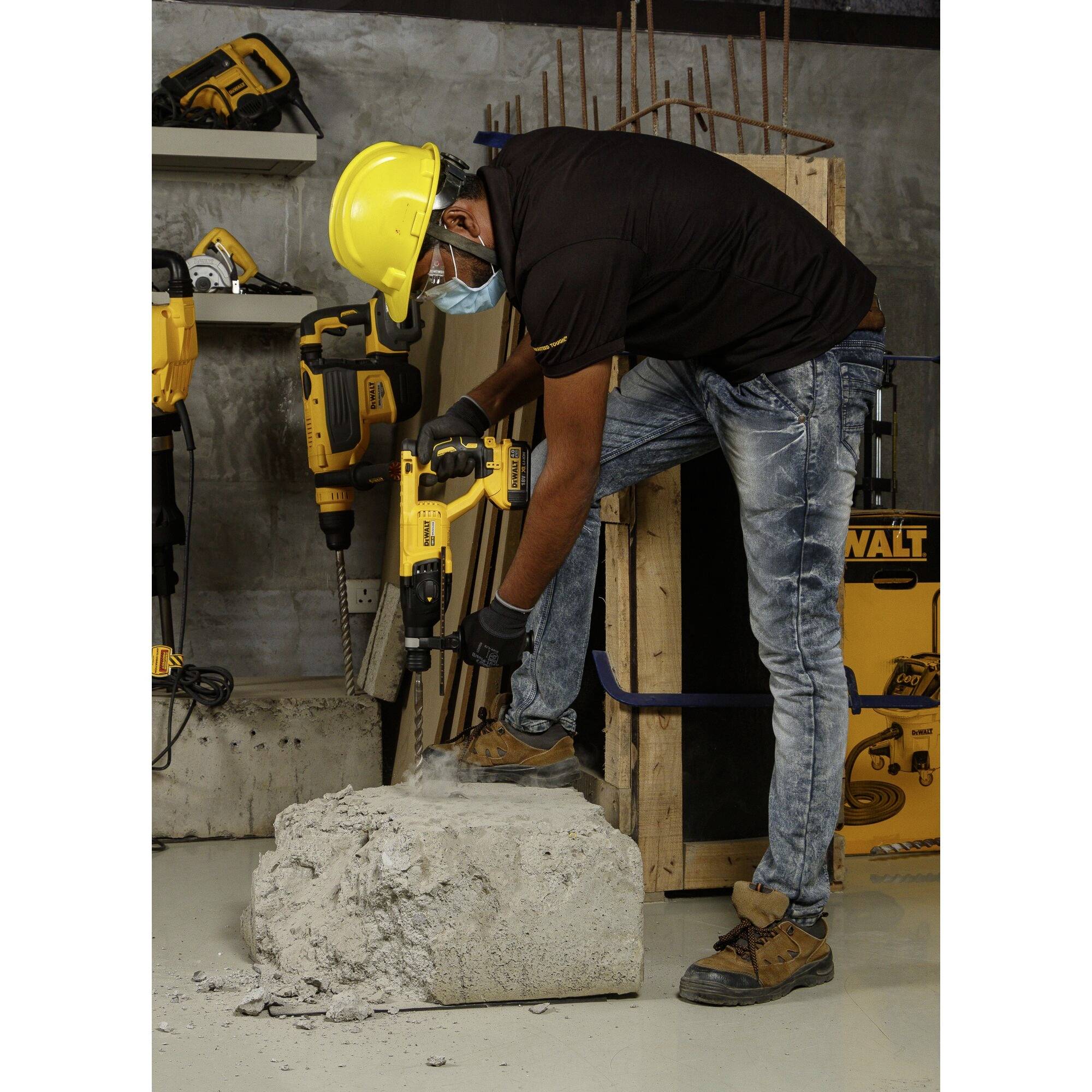Ein Bauarbeiter mit Schutzhelm und Maske bohrt mit einem gelben Bohrhammer in einen großen Betonblock. Im Hintergrund sind Werkzeuge.