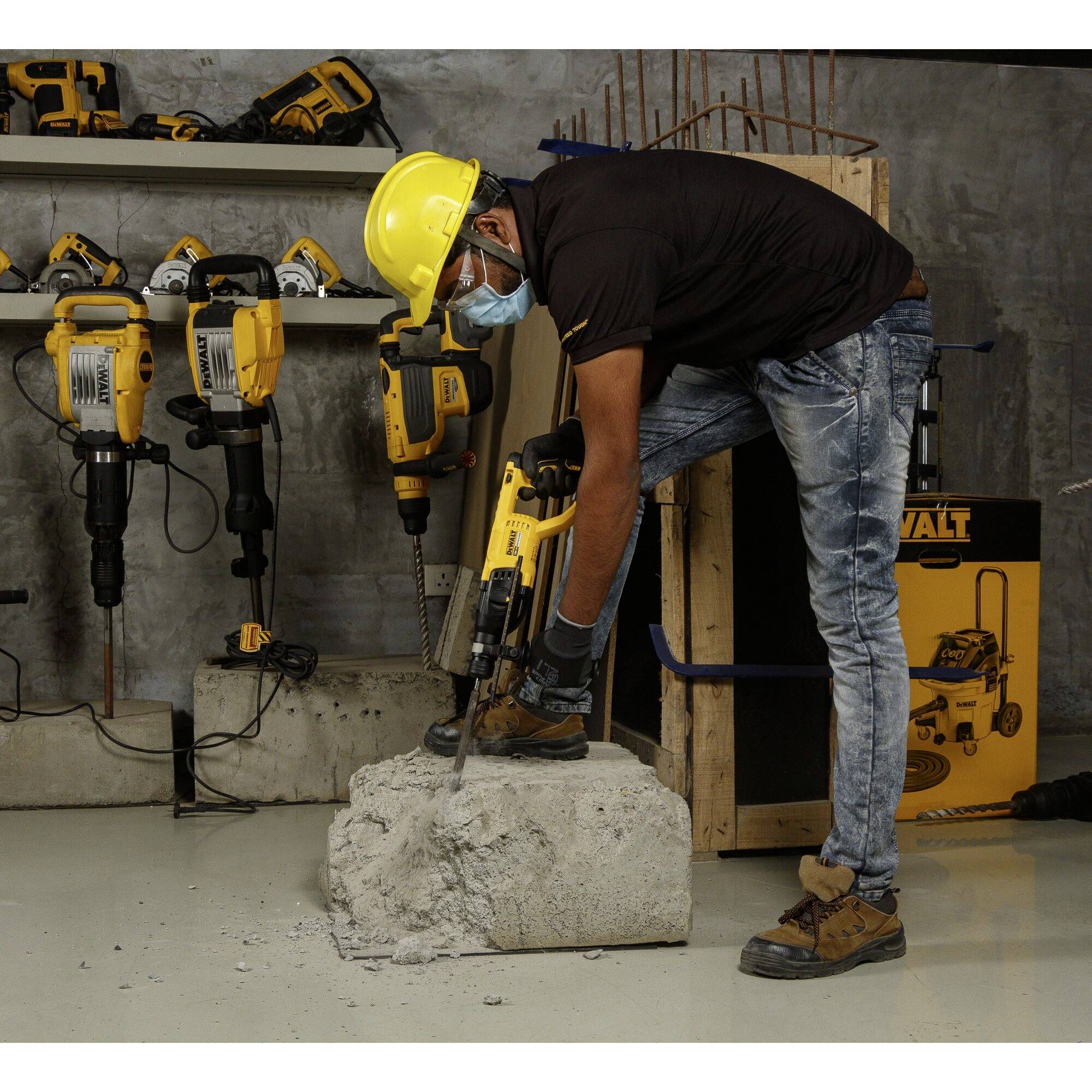 Ein Mann mit Helm und Maske bohrt mit einem Elektrowerkzeug in einen Betonblock. Im Hintergrund sind weitere Bohrgeräte zu sehen.