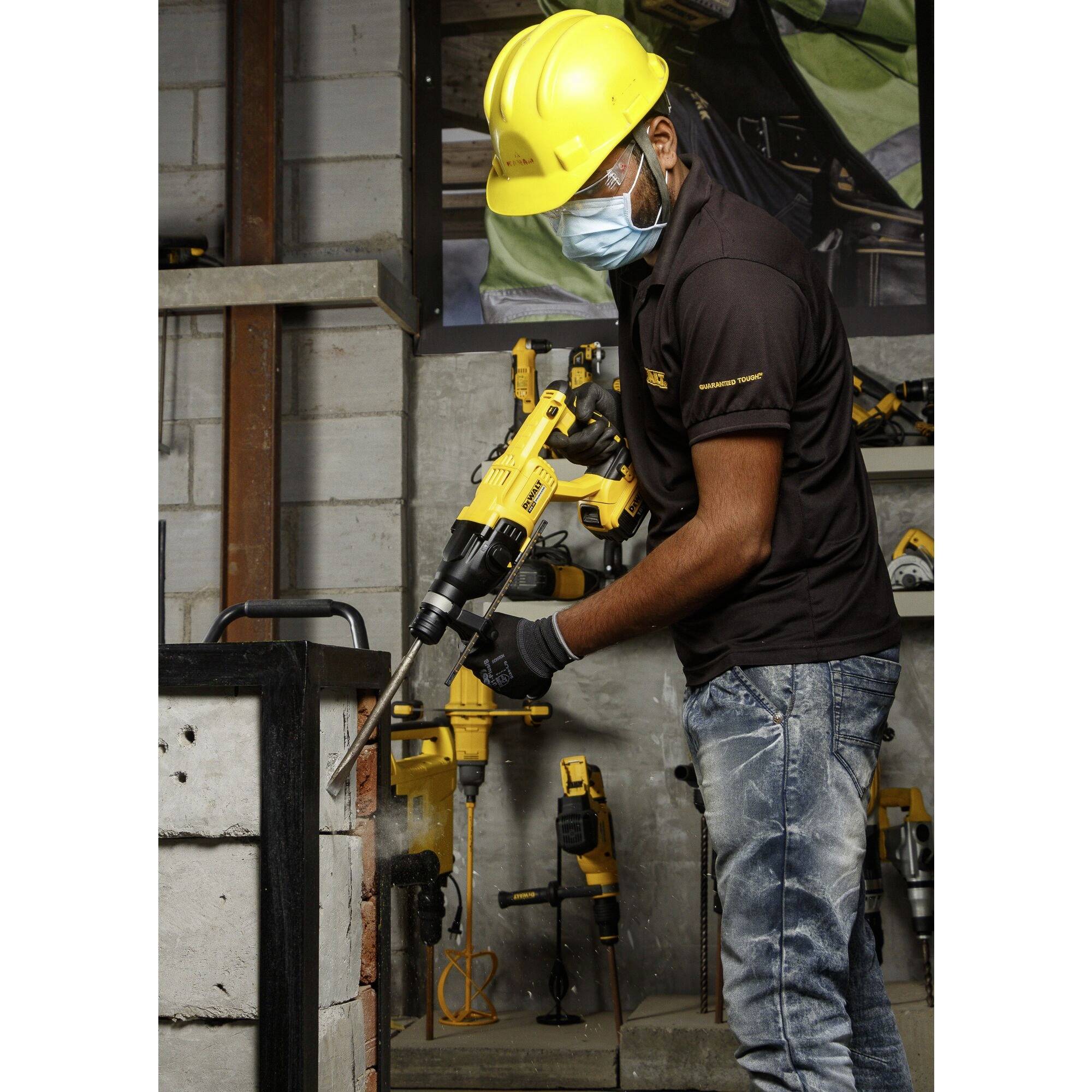 Eine Person mit Schutzhelm und Maske arbeitet mit einem Bohrhammer an einer Wand in einer Werkstattumgebung.