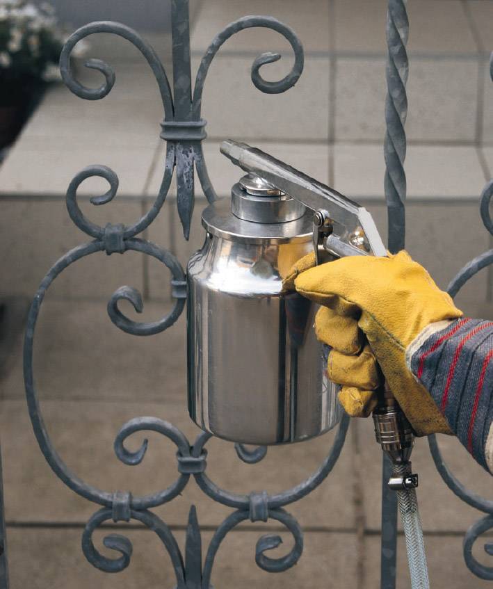 Person mit gelben Handschuhen lackiert ein Metallgitter mit einer silbernen Spritzpistole.