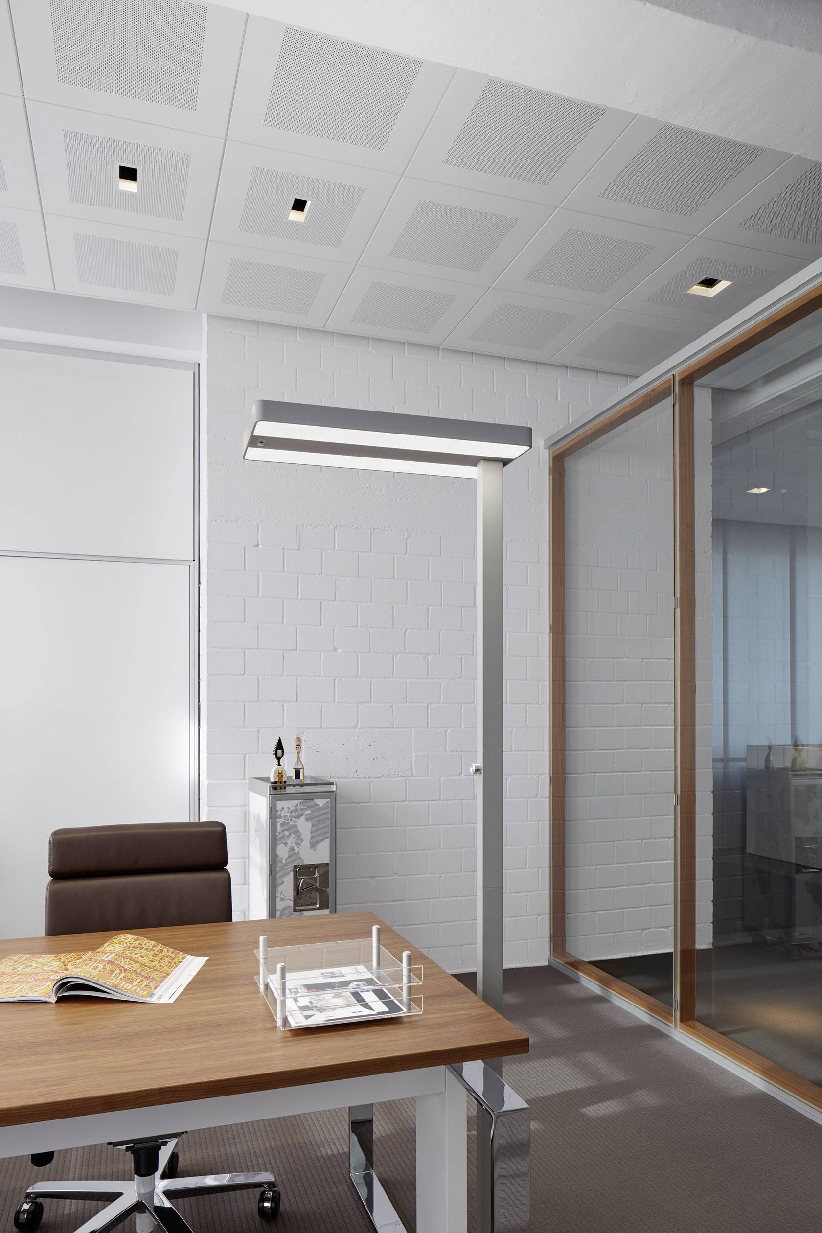 Ein modernes Büro mit einem Holztisch, einem offenen Magazin, einem braunen Stuhl, einer Lampe und einer weißen Backsteinmauer im Hintergrund.