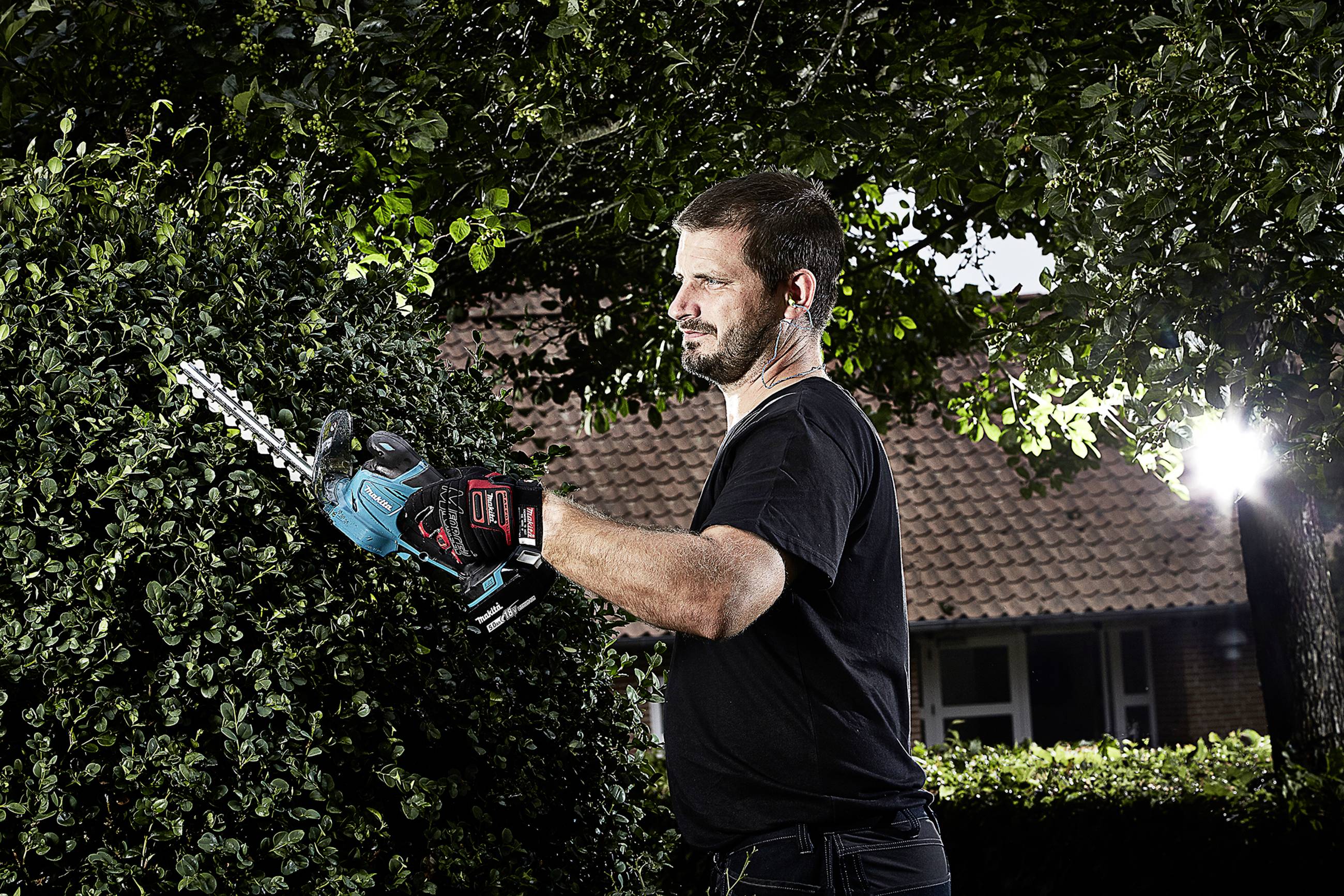 Mann schneidet mit einer elektrischen Heckenschere eine grüne Hecke, umgeben von Bäumen. Im Hintergrund ein Ziegelsteingebäude.