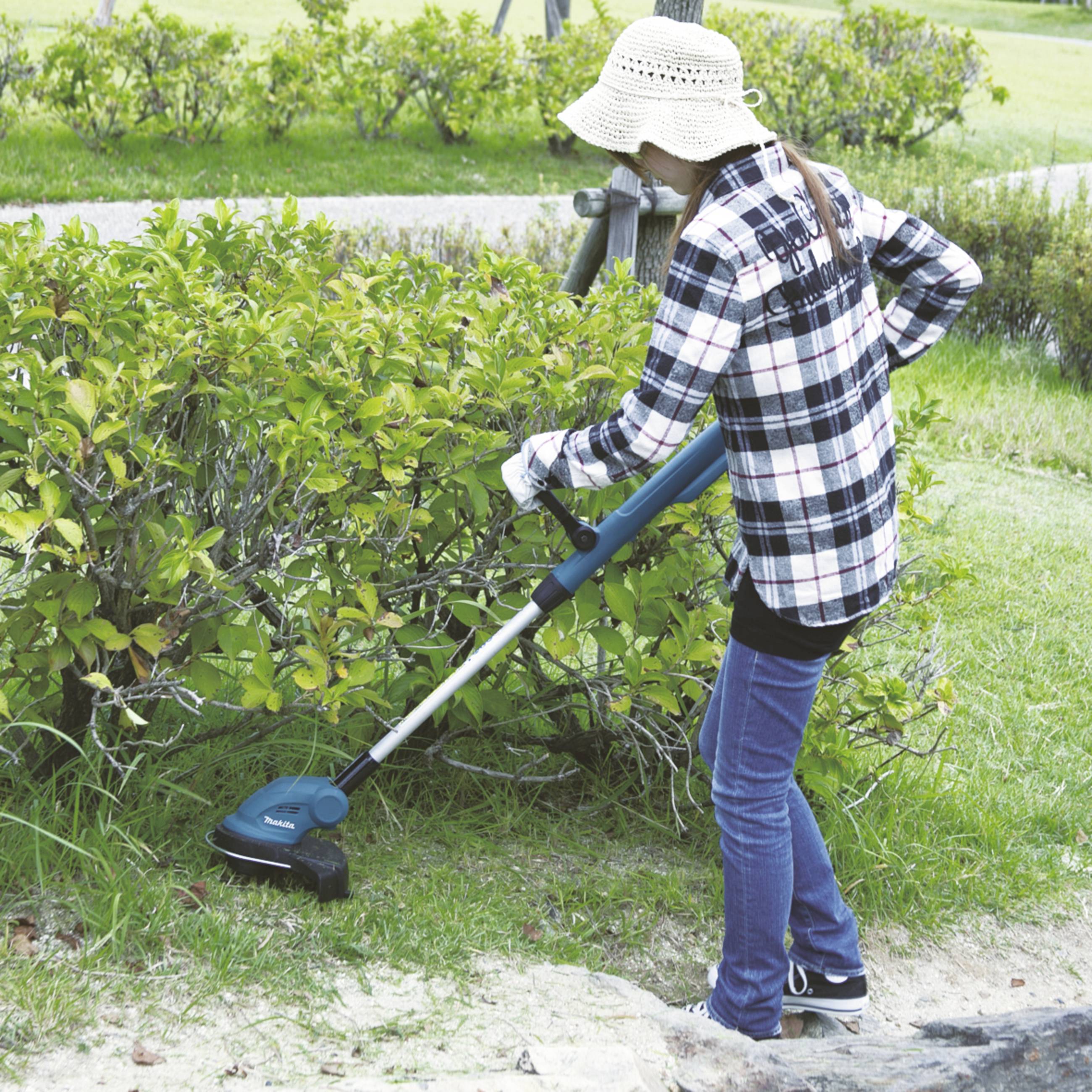 Eine Person mit Sonnenhut und kariertem Hemd schneidet Hecken mit einem elektrischen Trimmer in einem Garten.