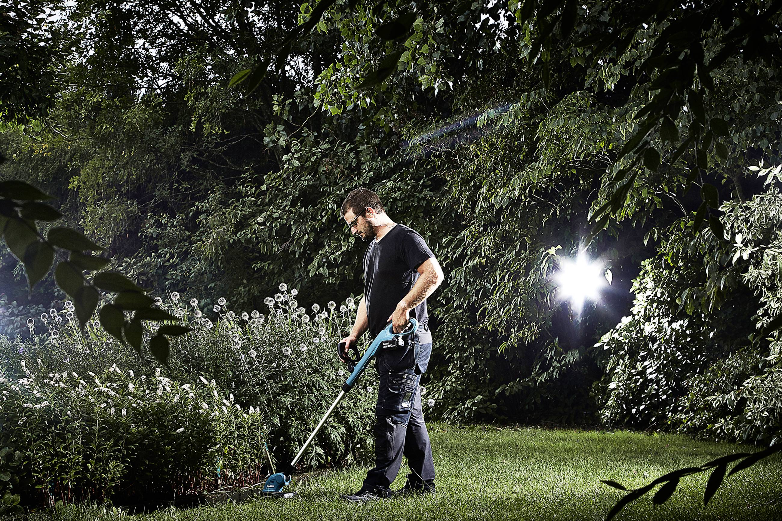 Eine Person benutzt einen Rasentrimmer im Garten, umgeben von Bäumen und Pflanzen. Es ist sonnig, wodurch Schatten auf den Rasen fallen.